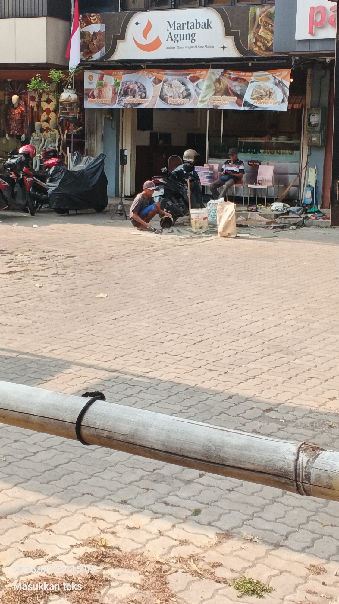 A street scene in Malang, Indonesia, shows a person crouching on a paved walkway in front of a building with a "Martabak Agung" sign. The person is wearing a cap, t-shirt, and shorts and appears to be working with a bucket and some materials on the ground. Two motorcycles are parked nearby, one covered with a black tarp. A man is seated on a chair further back in front of the building, looking at his phone. The building's facade features images of food items and the Indonesian flag. The environment suggests an urban street with shops or food stalls.