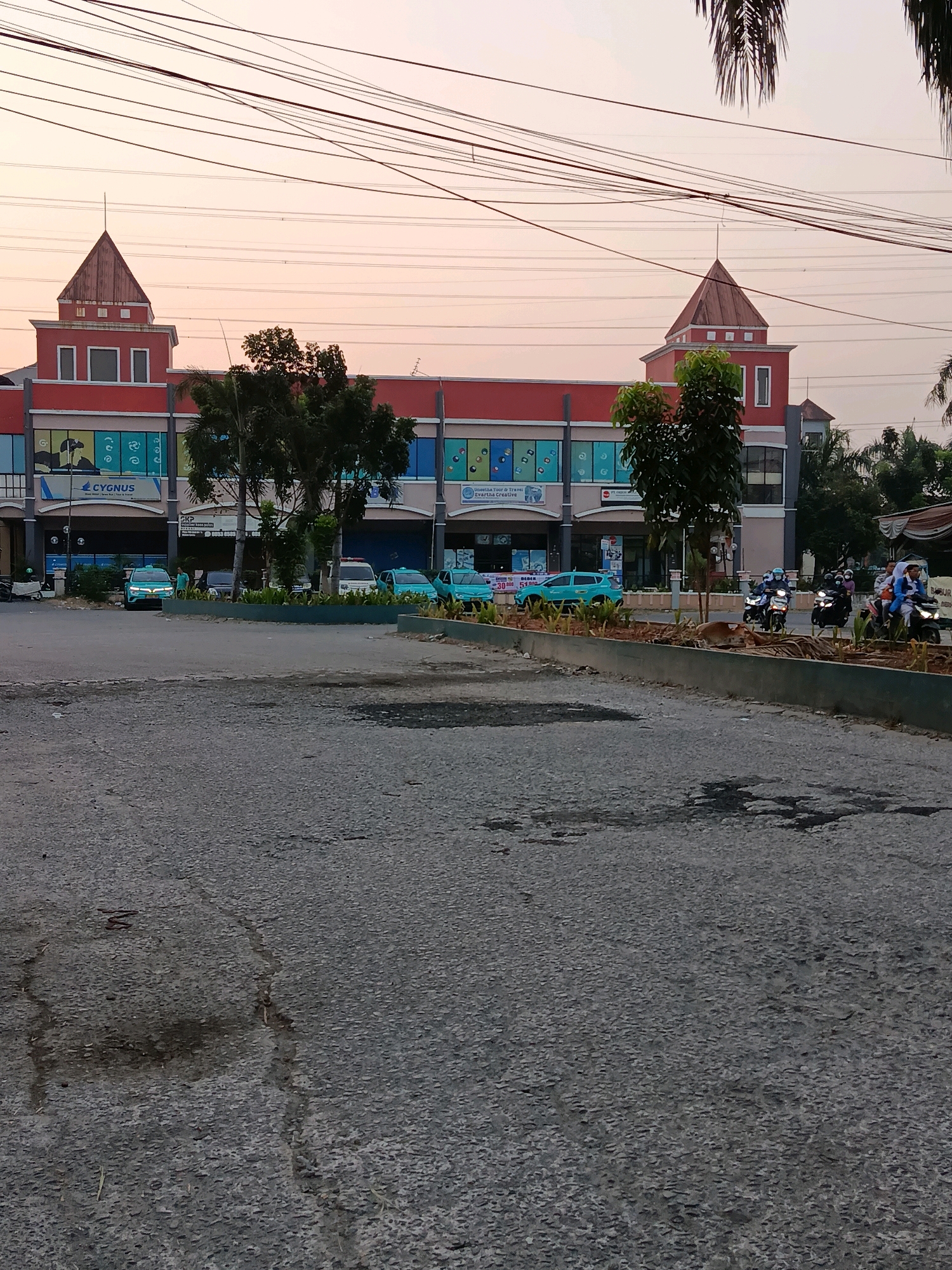 In Bekasi, Indonesia, a street scene unfolds during what appears to be dusk. A long, two-story building with a red facade dominates the background, featuring architectural elements reminiscent of a castle with two prominent spired towers. The building houses various storefronts, some with colorful signage and windows displaying graphics. Several turquoise vehicles, possibly taxis or ride-sharing cars, are parked and in motion on the street in front of the building. A herd of cattle is visible lying on the grass in the foreground, near a section of decorative landscaping. Motorcyclists are also present on the street, some carrying passengers. Overhead, a network of power lines crisscrosses the sky, indicating the presence of urban infrastructure. The lighting suggests either early morning or late evening, with a soft, diffused light illuminating the scene.