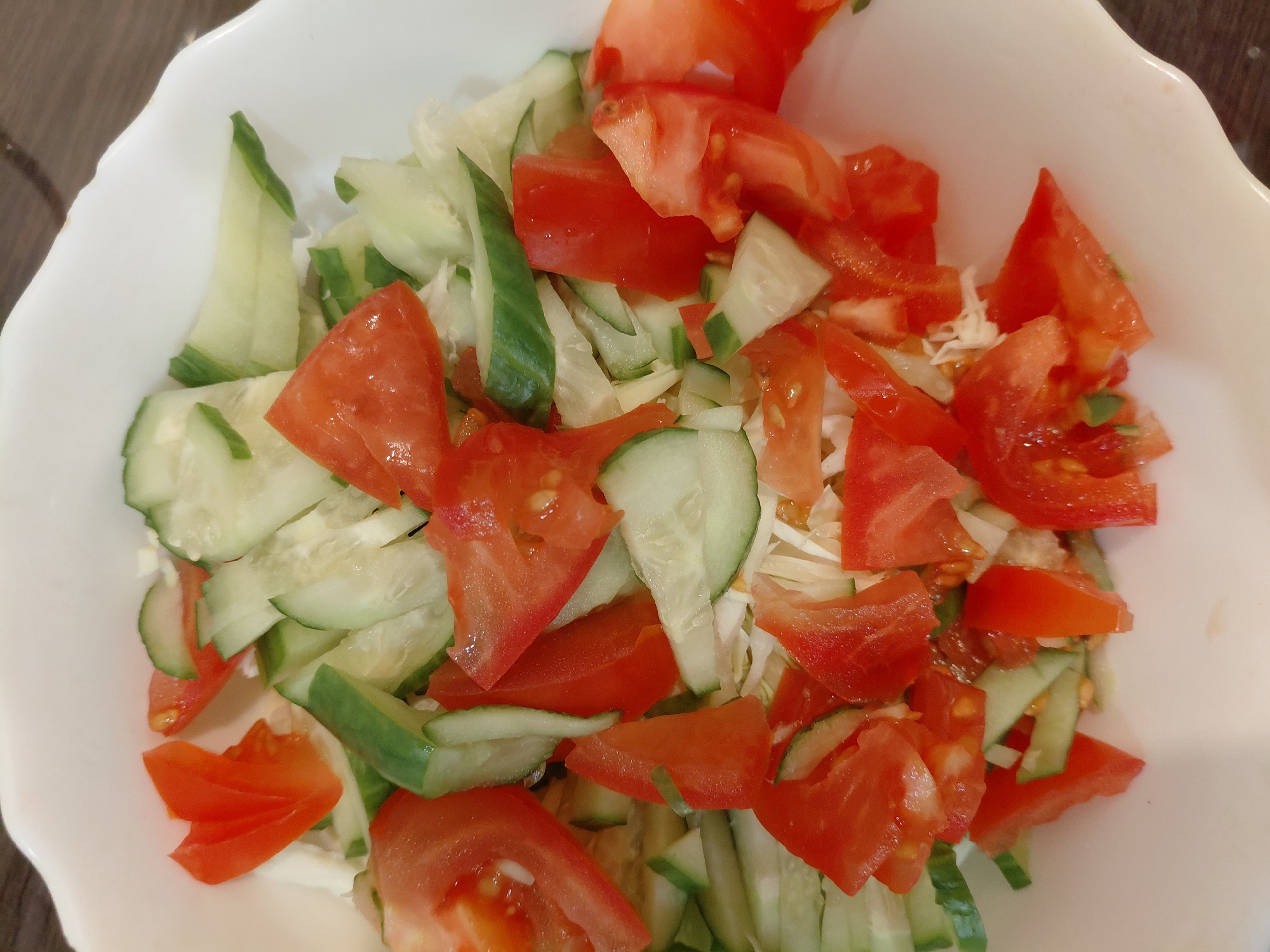 A close-up, top-down view shows a fresh vegetable salad presented in a white bowl with a fluted or scalloped rim. The salad is composed primarily of vibrant red chopped tomatoes and green sliced cucumbers. Interspersed among these are lighter, finely shredded or sliced white vegetables, likely cabbage or onion. The tomatoes are cut into irregular wedges, revealing some of their moist interior, while the cucumbers are sliced, displaying their green skin and lighter flesh. The ingredients are mixed together, filling the white bowl. The background surrounding the bowl is dark and out of focus, suggesting a table surface in an indoor setting. There are no people visible, nor any specific activity taking place other than the static presentation of the prepared salad. No text is visible in the image. The lighting is bright and even.