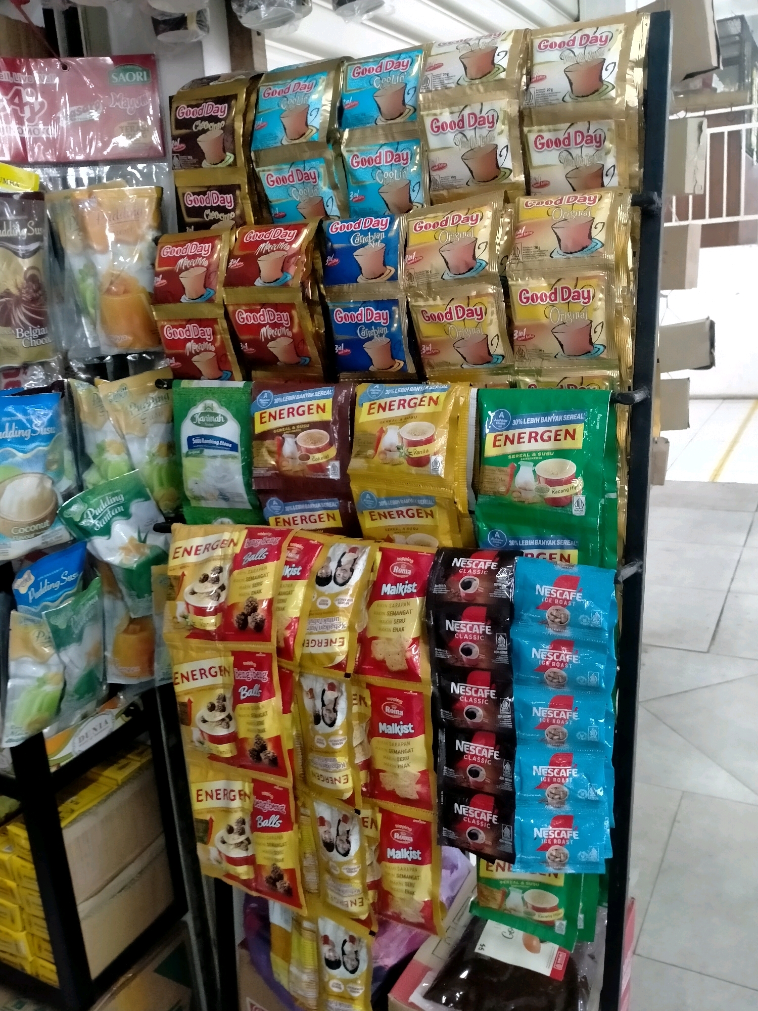 The image shows a retail display of various Indonesian food and beverage products. The display is organized on a black metal rack, with many items hanging and stacked. Prominently featured are sachets of instant coffee, notably "Good Day" and "Nescafe Classic" brands, as well as "Nescafe Ice Roast." There are also bags of "Energen" cereal and milk drinks, "Pudding Susu" (milk pudding), and "Pudding Santan" (coconut milk pudding). Some items are in larger packaging, such as "Saori" seasoning and "Roma Malkist" crackers. The background suggests an indoor retail environment, possibly a small warung or minimarket, with other goods and shelving visible. The overall impression is a well-stocked display intended for casual consumer purchase. The presence of these specific brands and product types indicates the location is likely in Indonesia, given their widespread availability there. Batu, a city in East Java, Indonesia, is known for its agricultural products and tourism, and this type of local convenience store or market is common throughout the region.