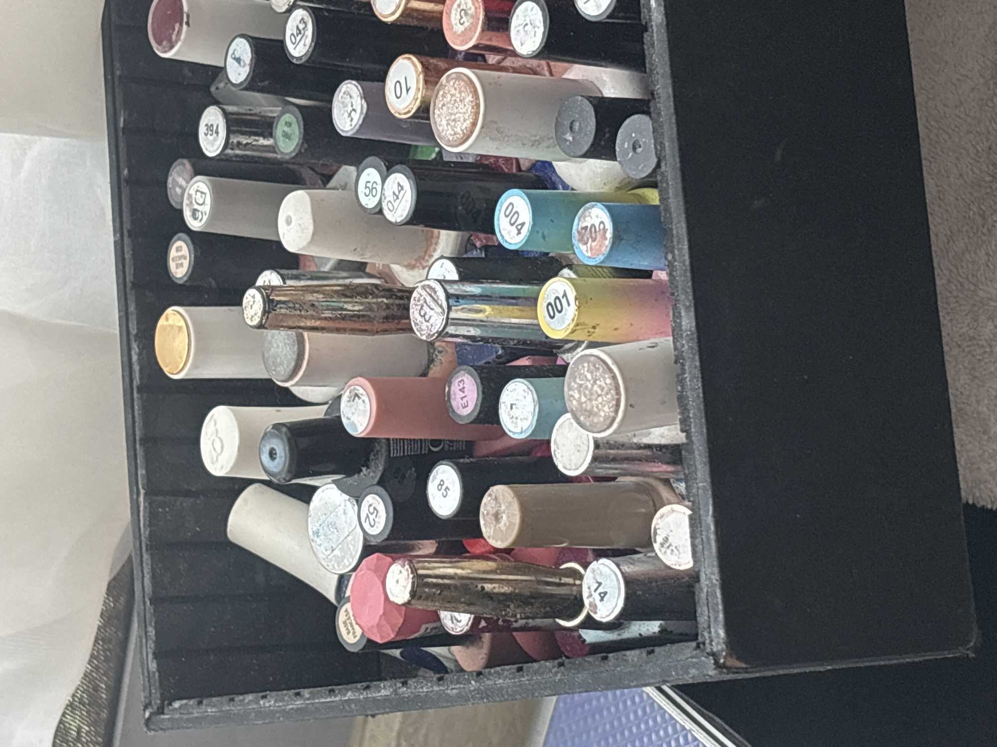 A black storage container, possibly a drawer or box, is filled with numerous small, cylindrical bottles, likely nail or gel polishes, in Jesús del Monte, Mexico. The bottles are arranged horizontally, creating a dense, somewhat disorganized collection. Their caps display a wide array of colors and finishes, including metallic, glitter, solid, and translucent shades of red, pink, gold, silver, black, white, cream, blue, teal, and purple.
Many caps feature circular labels with numbers and some text. Clearly visible numbers include 043, 394, D6, 56, 044, 004, 709, 001, E143, 85, 52, 14, and 10. Text visible on specific caps includes "BASE FRANCESA" on a pink octagonal cap and "RED FRANCESA" on another. The background shows hints of a white fabric on the left and a darker, textured surface on the right. No people are present, and the bright, even lighting suggests daytime.