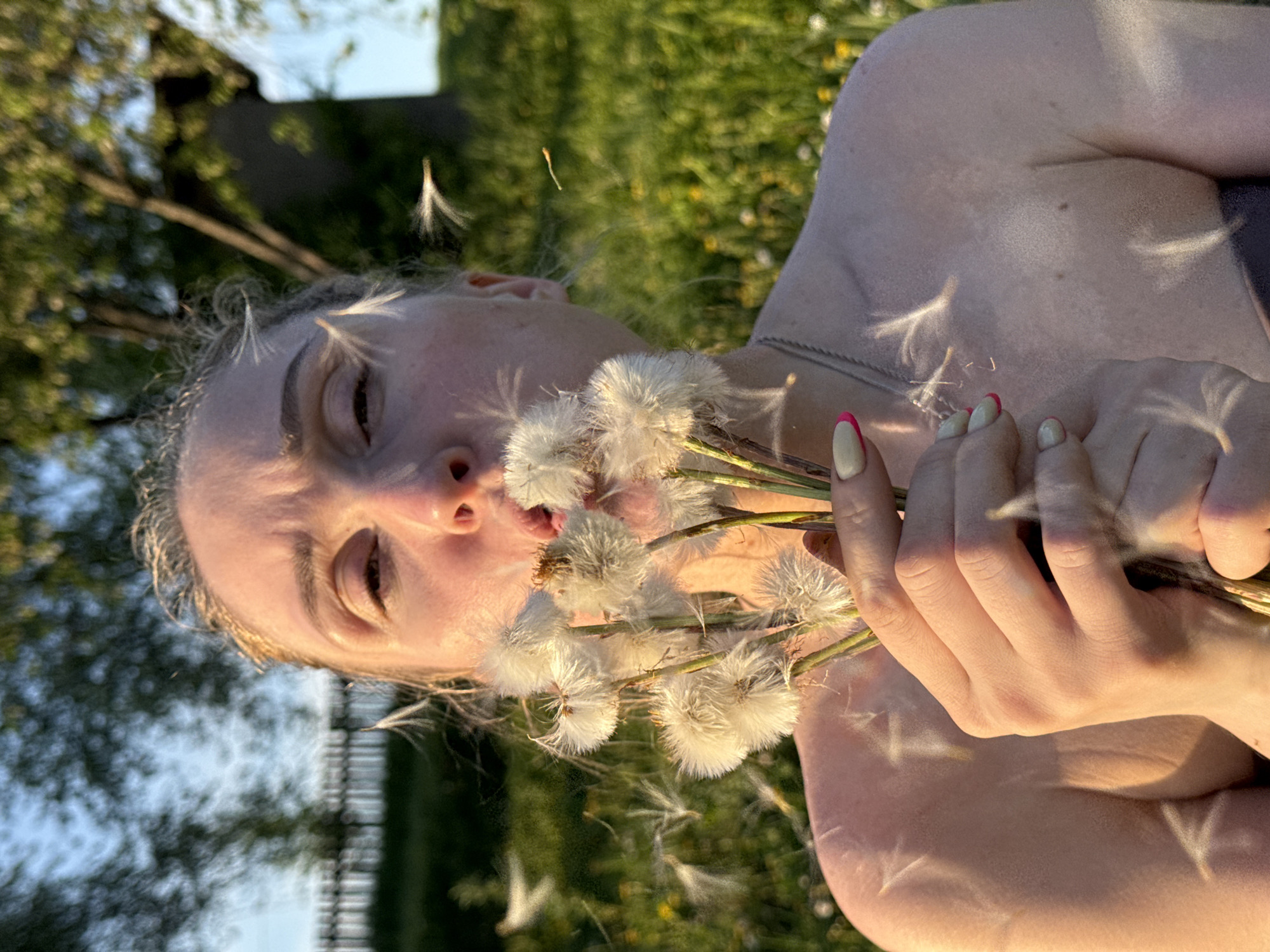 A fair-skinned woman is holding a bunch of white, fluffy dandelions up to her face, blowing the seeds off in an outdoor setting. She has light-colored hair, and her nails are painted with light colors with red accents. She is wearing what appears to be a dark top with thin straps. The background includes green foliage and a blurred building, suggesting an outdoor location. The presence of dandelions suggests it is springtime. There is no evidence that this is in Nizhnyaya Chermoda, Russia.