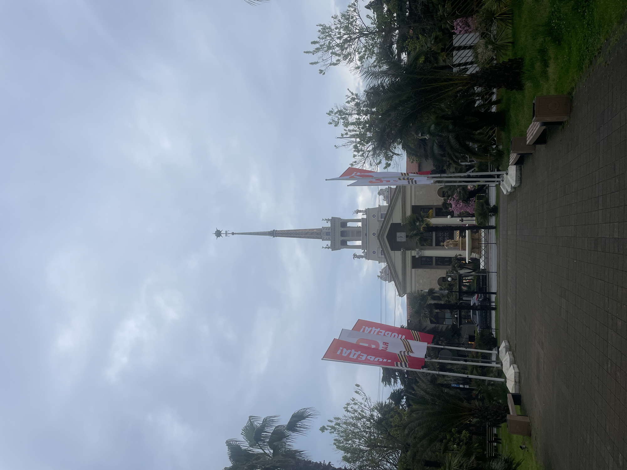 The image captures a view of the Sochi Sea Port building in Sochi, Russia. The building features a prominent spire and classical architectural details. Several flags displaying the phrase "ПОБЕДА!" (Victory!) are in the foreground. The sky is overcast. The surrounding area includes palm trees, other vegetation, and a paved walkway with benches. No people are clearly visible.