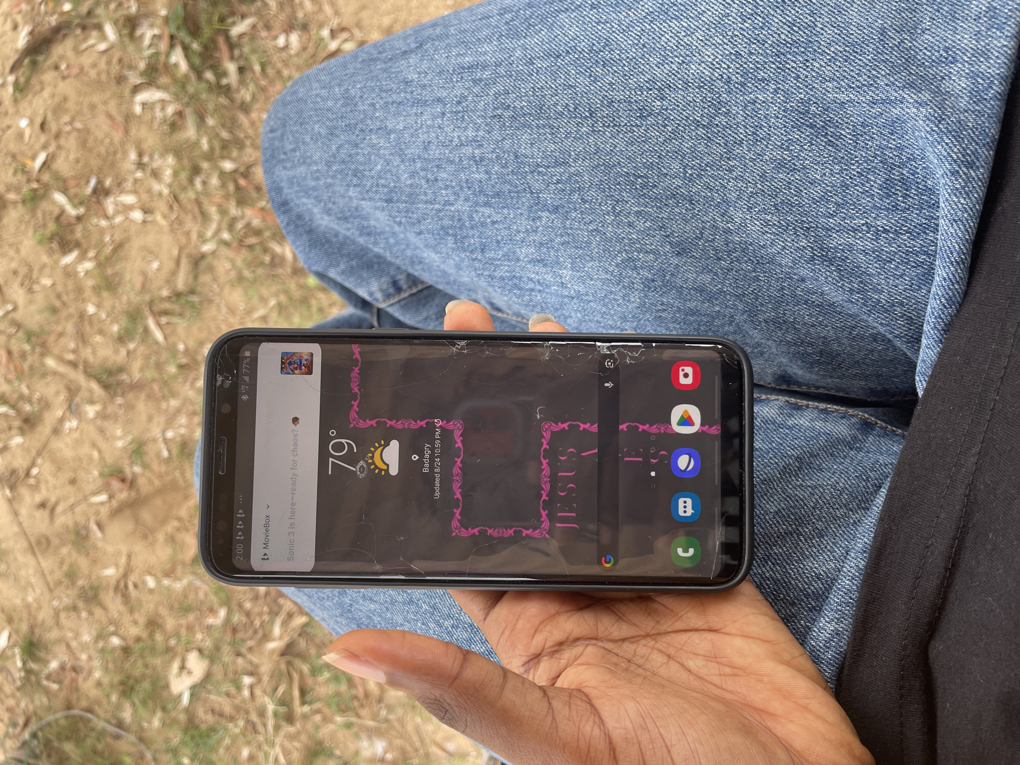 A person holds a cracked smartphone displaying a weather widget showing 79 degrees and a cloudy sky. The phone's wallpaper features pink ornate borders and the word "JESUS" in stylized lettering. The user's left hand, with short fingernails, is visible gripping the phone. The background consists of dry ground with scattered leaves and twigs. The person is wearing blue denim jeans and a black shirt. The image was taken in Iyafin, Nigeria.
