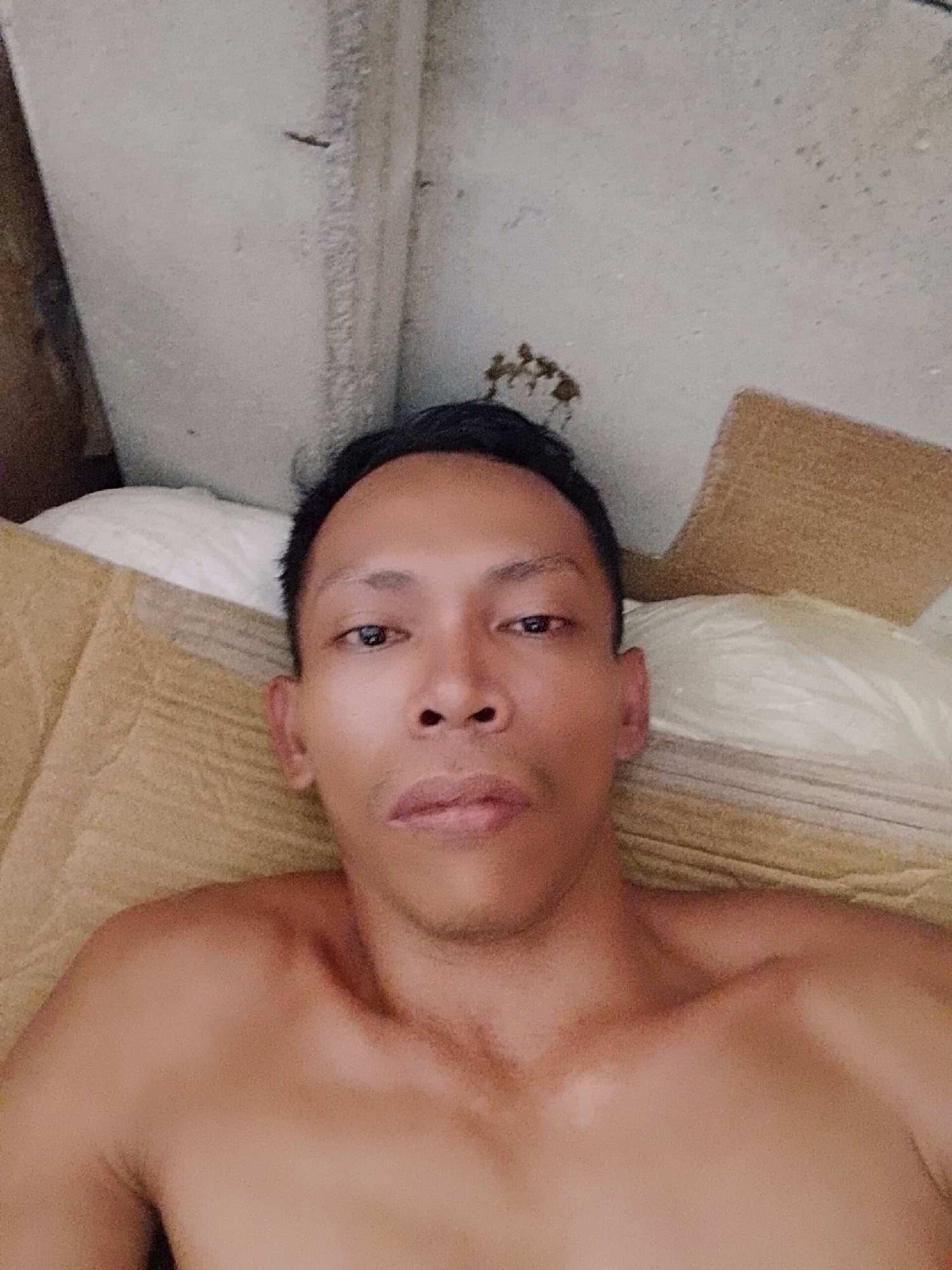A shirtless man is lying down, possibly on a bed, in an indoor environment. The man is looking directly at the camera. A pillow and blanket are partially visible. The background shows an unfinished concrete wall and a brown object, potentially cardboard. The location is Bukit Ketok, Indonesia.