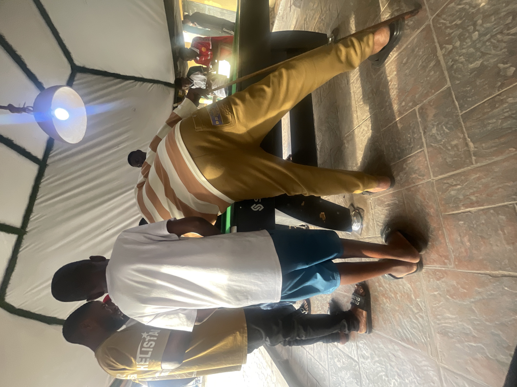 Three individuals are observed around a pool table beneath a light fixture inside a tent-like structure. The location is identifiable as Lokoja, Nigeria, due to the presence of a patch with the letters "PP" possibly indicating the region in Nigeria. One person wears tan trousers and a striped shirt, holding a pool cue. Another wears a white shirt and blue shorts. A third person wears a mustard-colored shirt with the word "CHELISTA" on it and black trousers. All three are wearing sandals.
