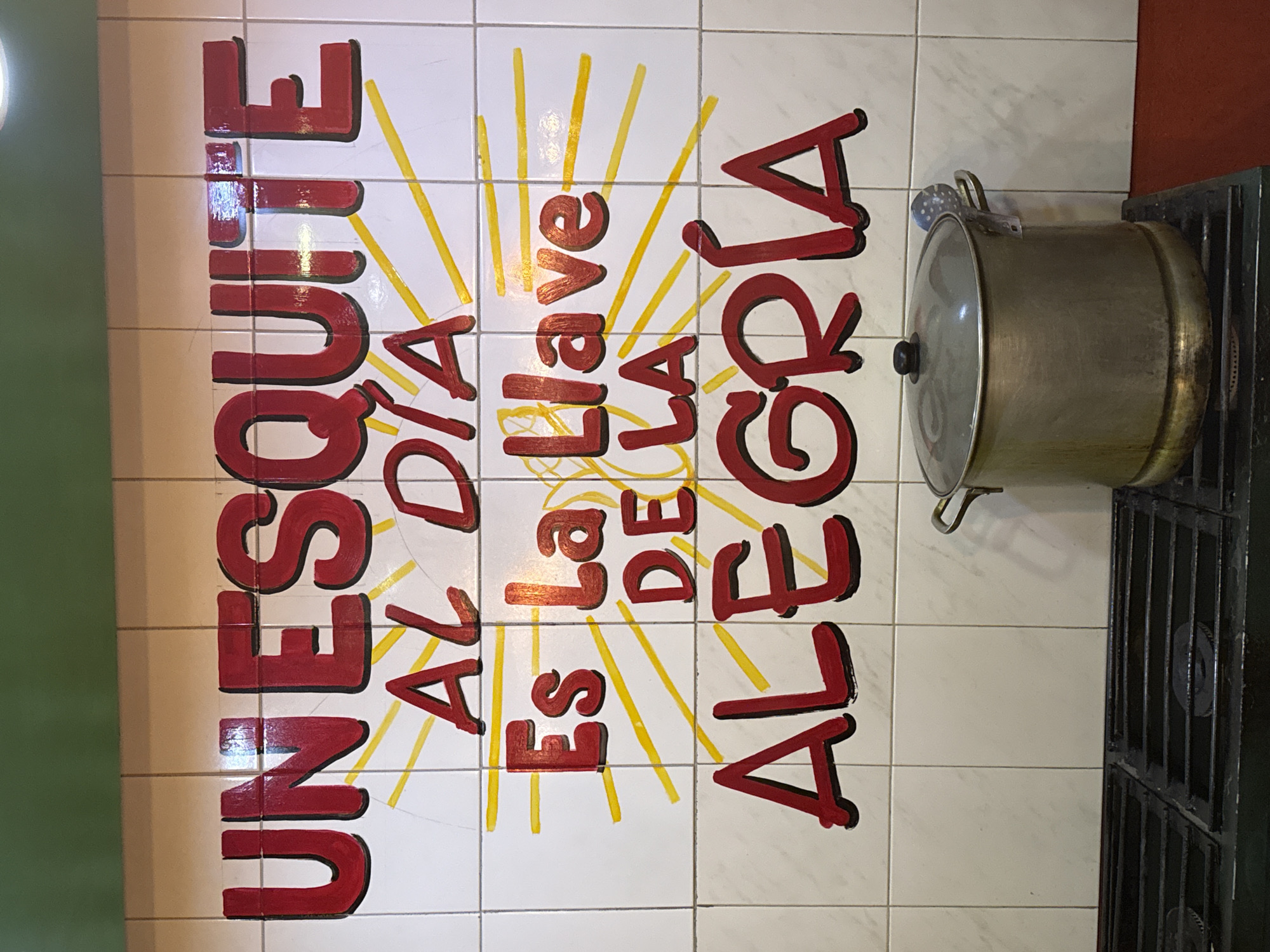 An indoor scene in San Bernardino Tlaxcalancingo, Mexico, displays a tiled wall above a commercial-style gas stove. The wall consists of white square tiles, approximately 15x15 cm, with visible grout lines and a green-painted section above. Dominating the wall is hand-painted text in bold red letters with black outlines, which reads: "UN ESQUITE AL DÍA Es La Llave DE LA ALEGRÍA" (An Esquite a Day Is The Key To Happiness). Yellow painted lines, suggesting sun rays, emanate from behind sections of the text. Below the wall, a black, multi-burner gas stove features a large, metallic pot with a lid resting on its rightmost grate. A metal strainer spoon is hooked onto the pot's visible right handle. No individuals are present in the static scene.