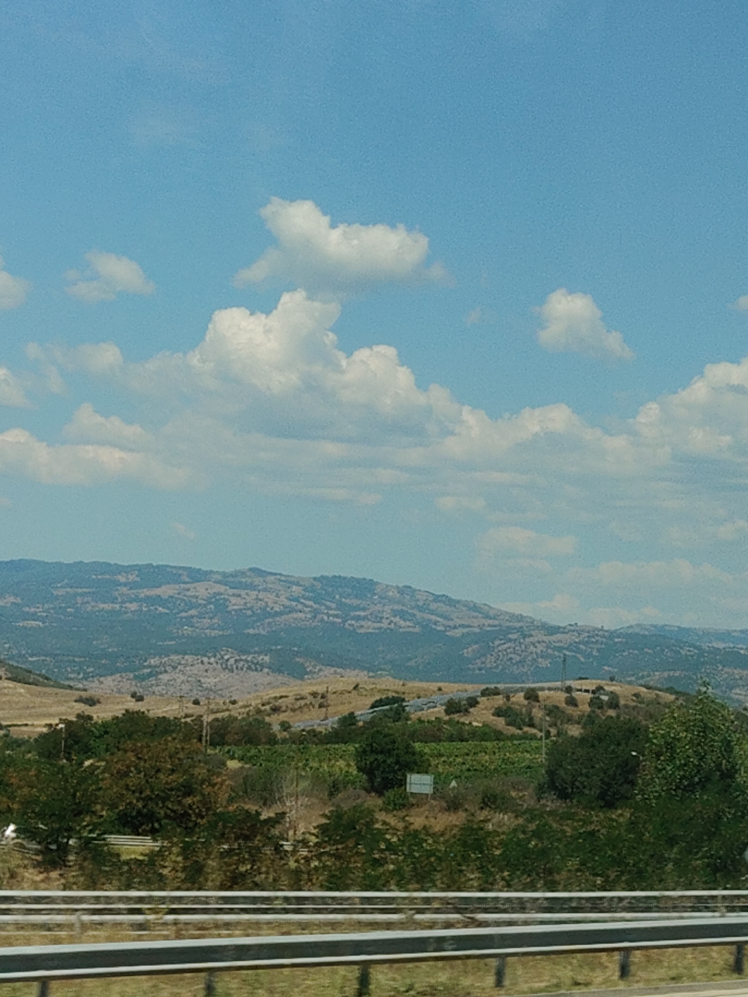 The image displays a serene, rural landscape under a bright blue sky dotted with fluffy white clouds. The foreground features a highway barrier, suggesting a view from a moving vehicle. Beyond the barrier, a lush green expanse of trees and vegetation stretches across the middle ground, interspersed with cultivated fields. In the distance, rolling hills and mountains form a picturesque backdrop, their slopes covered in a mosaic of earthy tones and greenery. A few scattered structures, possibly farmhouses or rural buildings, are visible among the hills. The scene evokes a sense of tranquility and natural beauty, characteristic of the Bulgarian countryside, potentially near the village of Levunovo, known for its scenic surroundings.
