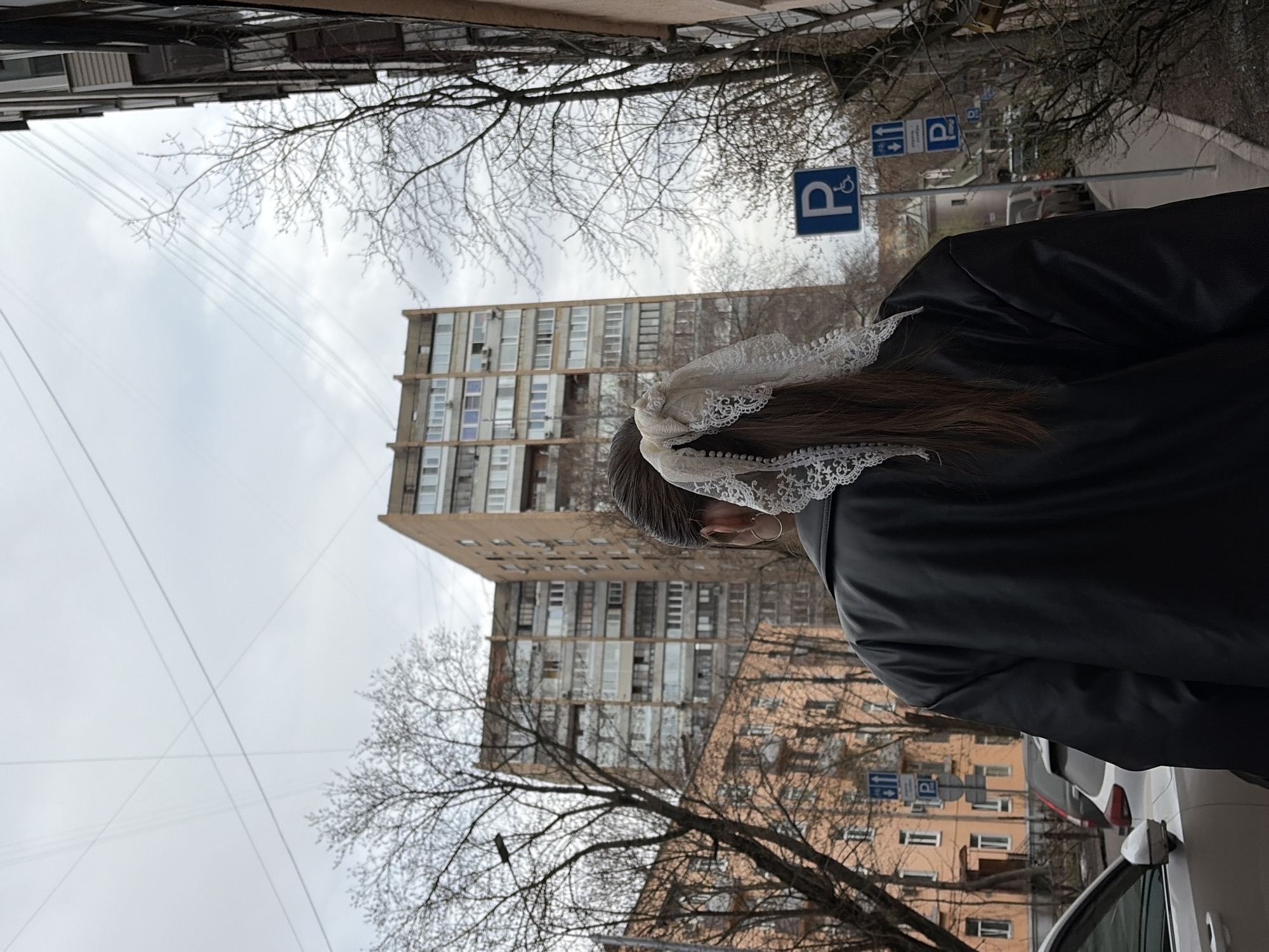 A low-angle, outdoor shot shows a person wearing a black leather jacket and a decorative white lace hair accessory. In the background, there are multi-story buildings, bare trees, street signs indicating disabled parking, and overhead power lines against a cloudy sky. The buildings suggest an urban environment, possibly Moscow, Russia. A white car is partially visible in the foreground to the right.