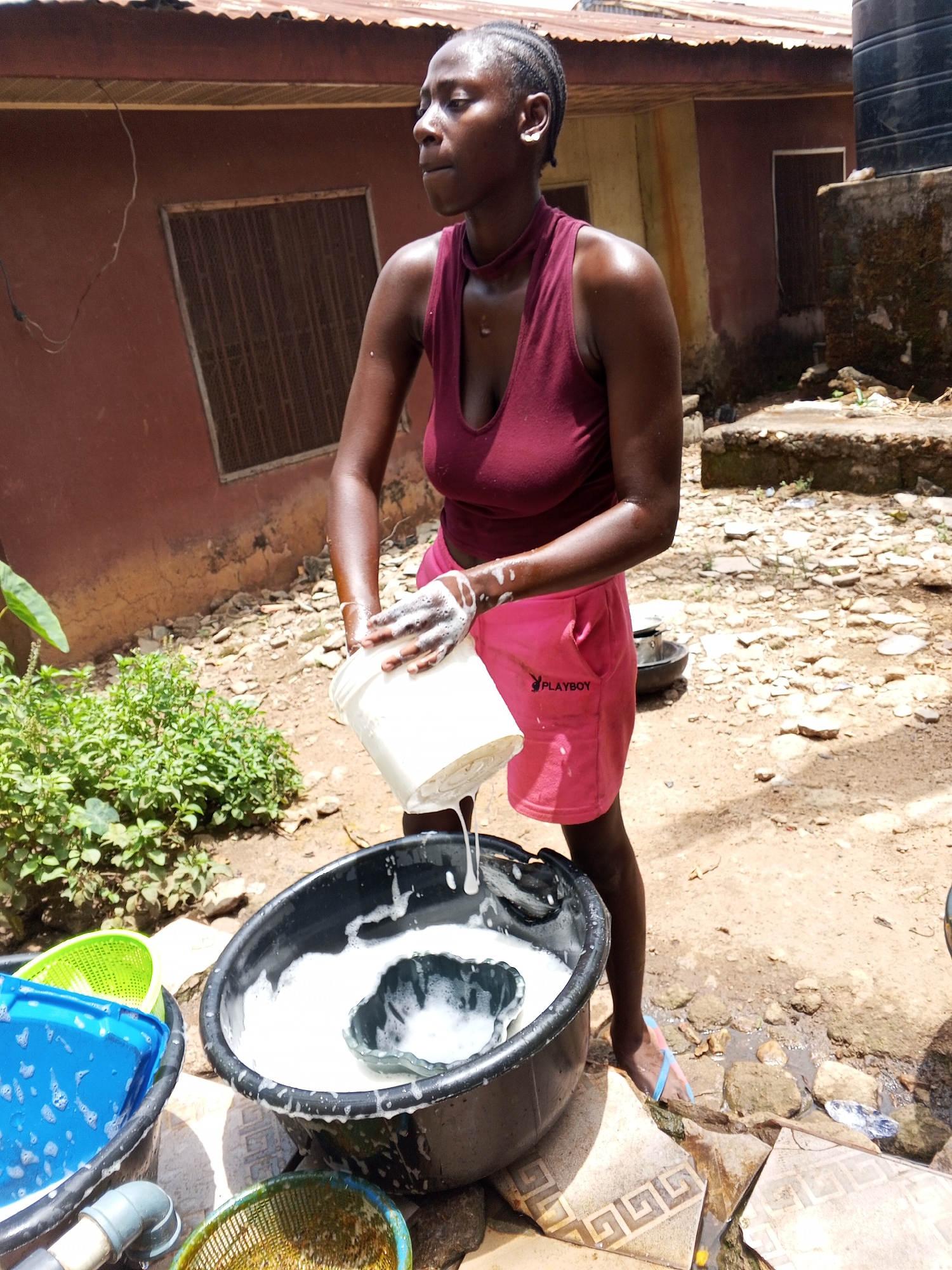 A woman is standing in a yard outside a reddish-brown building washing laundry. She is pouring water from a white bucket into a black tub filled with soapy water. The woman is wearing a maroon tank top and pink shorts with "PLAYBOY" printed on them. There are other tubs and containers around the black tub, filled with soapy water or other contents. The yard is dirt and stone, with some vegetation. The building has a window with a barred screen. The exact location in the city of Akwanga, Nigeria is not discernible from the image.