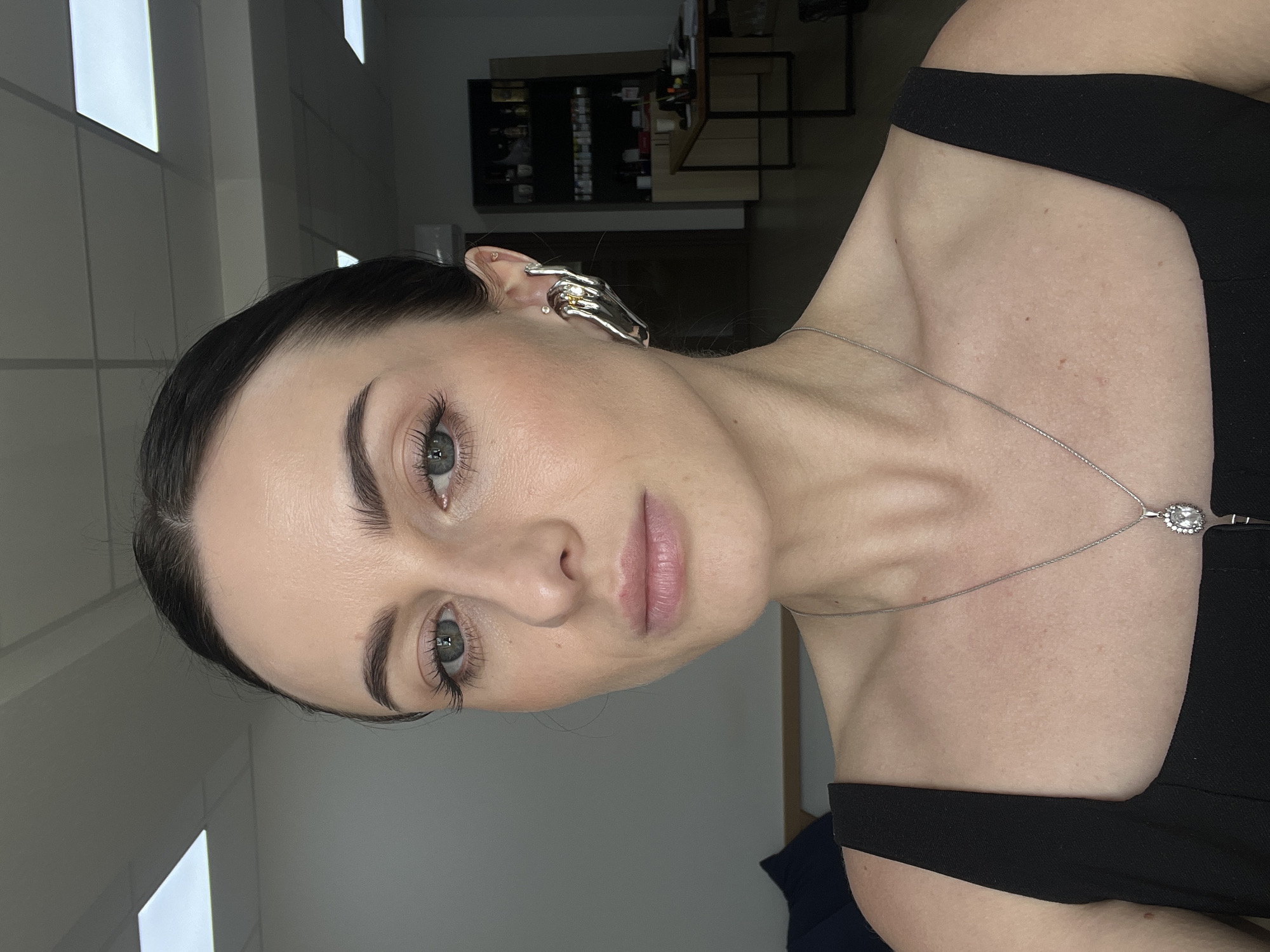 A medium shot portrait features a woman with dark hair pulled back, looking directly at the camera. She has light skin, green eyes, and prominent eyelashes. She is wearing a black top with a square neckline and a silver necklace with a pendant. A large, statement earring in the shape of a hand is worn on her right ear. The background is an indoor setting with fluorescent lighting on the ceiling, a wall, and office furniture including shelves with bottles and a desk with papers. The scene is devoid of any explicit Lviv, Ukraine identifiers.