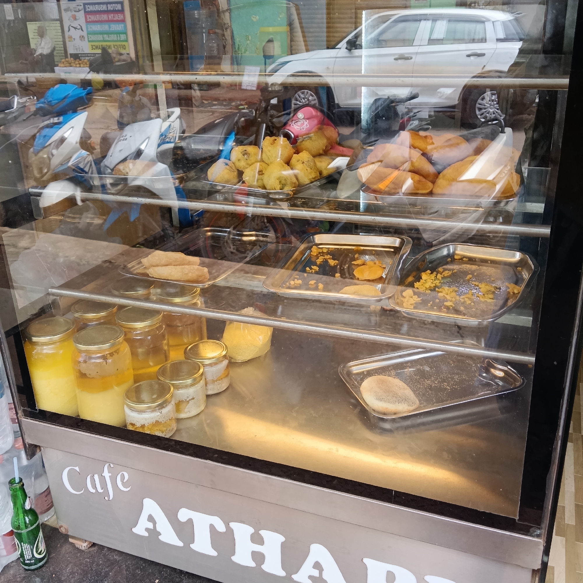 The image shows a display case from "Cafe Atharva", filled with assorted food items and jars of ingredients. On the top shelf are fried snacks, including what appear to be vada. The next shelf down contains trays with remnants of what seem to be fried items, alongside a tray of what may be papads. The bottom shelf holds several jars filled with yellow and white substances, likely ghee and lassi, and a tray with a single item. Outside the case are a few bottles, including a green glass bottle with a straw. Reflections in the glass reveal scooters, a white car, and other street elements. The location is in Oxel, India.