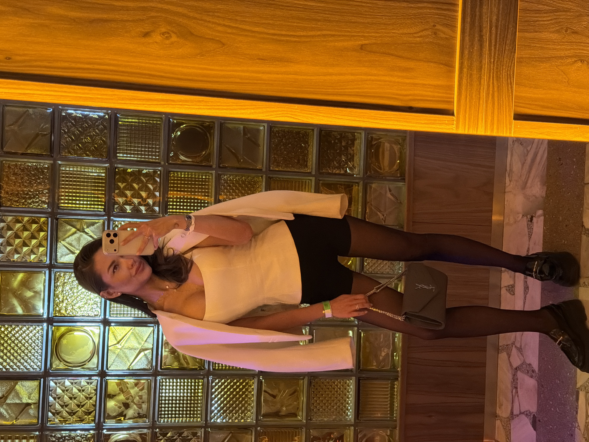 A young woman is captured in a full-length mirror selfie, posing upright against a distinctive interior background. She wears a white sleeveless top or dress, black short shorts or skirt, sheer black tights, and black chunky platform shoes or boots. A light-colored blazer or jacket is draped over her shoulders. Her left wrist displays a metallic watch, and her right wrist features a green and white band. She carries a small, grey shoulder bag with a gold chain strap, prominently featuring the "YSL" logo on its flap. She holds a light-colored smartphone to take the photo, showing a slight smile and direct gaze.
The background consists of a wall made of numerous square glass blocks, each featuring different textured patterns such as grids, circles, and abstract designs, reflecting warm, golden light. Above the glass block wall, a rich, amber-toned wooden structure, possibly a wall or ceiling panel, with visible grain, extends across the top of the frame. The floor below her appears to be a mixed material, with segments of light-colored, possibly marble or tiled, patterns interspersed with darker, granular sections. The overall lighting is warm and artificial, suggesting an indoor setting like a stylish restaurant, bar, or hotel lobby, likely during the evening.
