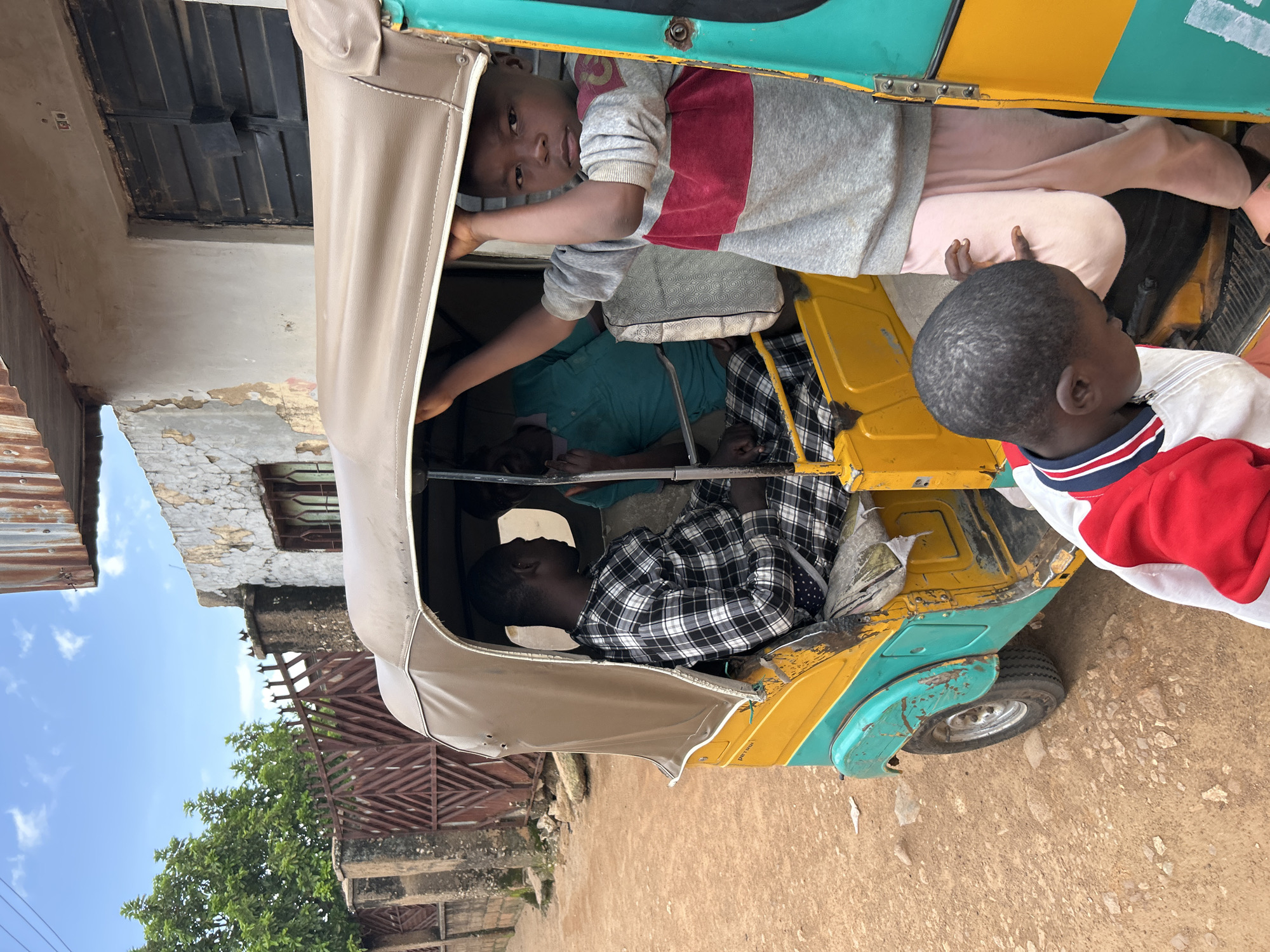 The image shows several children inside and around a yellow and turquoise auto-rickshaw, a common mode of transport in Jos, Nigeria. The auto-rickshaw is parked on a dirt road, with a building and some greenery visible in the background. The sun is shining, suggesting it is daytime, and the sky is clear blue.
In the foreground, a young boy with dark hair is looking towards the right of the frame. He is wearing a white shirt with red and blue trim. Another child, a girl, is visible inside the auto-rickshaw, leaning out and looking towards the camera. She is wearing a grey sweatshirt with a red stripe. Behind her, another child is lying down inside the rickshaw, wearing a blue shirt. Further inside, a child in a black and white checkered shirt is also visible.
The auto-rickshaw appears to be old and somewhat worn, with visible scratches and dents on its exterior. The building in the background is made of stone and has a corrugated metal roof. There is a brown metal gate and lush green trees.
The overall impression is a snapshot of everyday life in Jos, Nigeria, with children interacting with their surroundings and their mode of transportation. There is no visible text in the image.