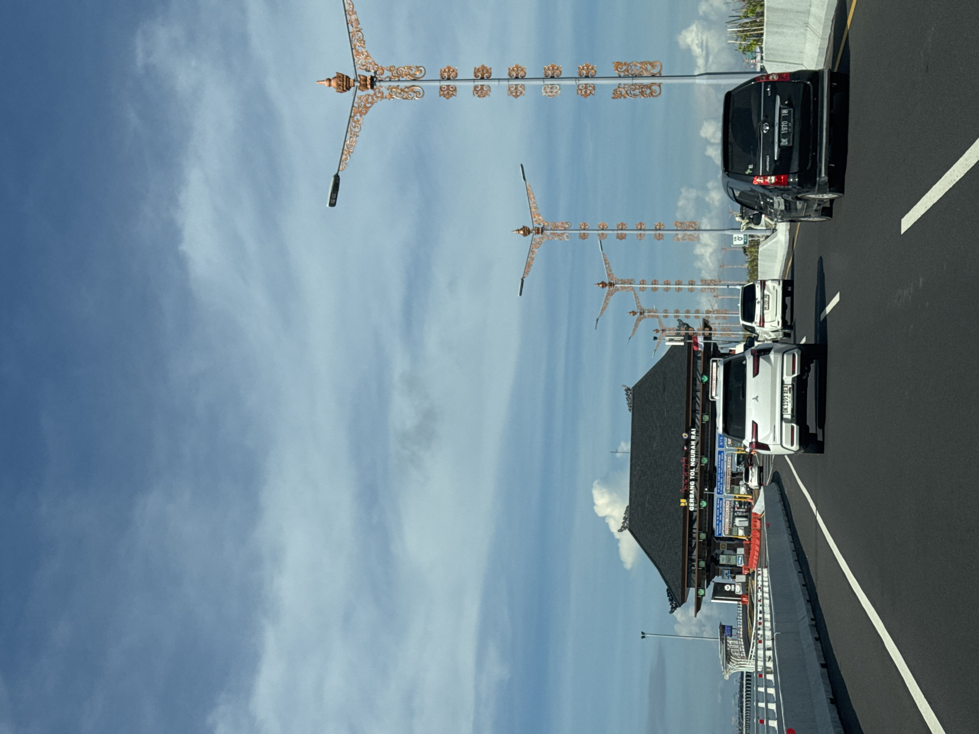 The image depicts a highway in Kuta, Indonesia, identifiable by the decorative streetlights and architecture. A toll booth labeled "GERBANG TOL NGURAH RAI" is visible. Several cars are present on the road, including a black van with license plate "DK 1970 IB" and a white car. The sky is blue with scattered clouds.