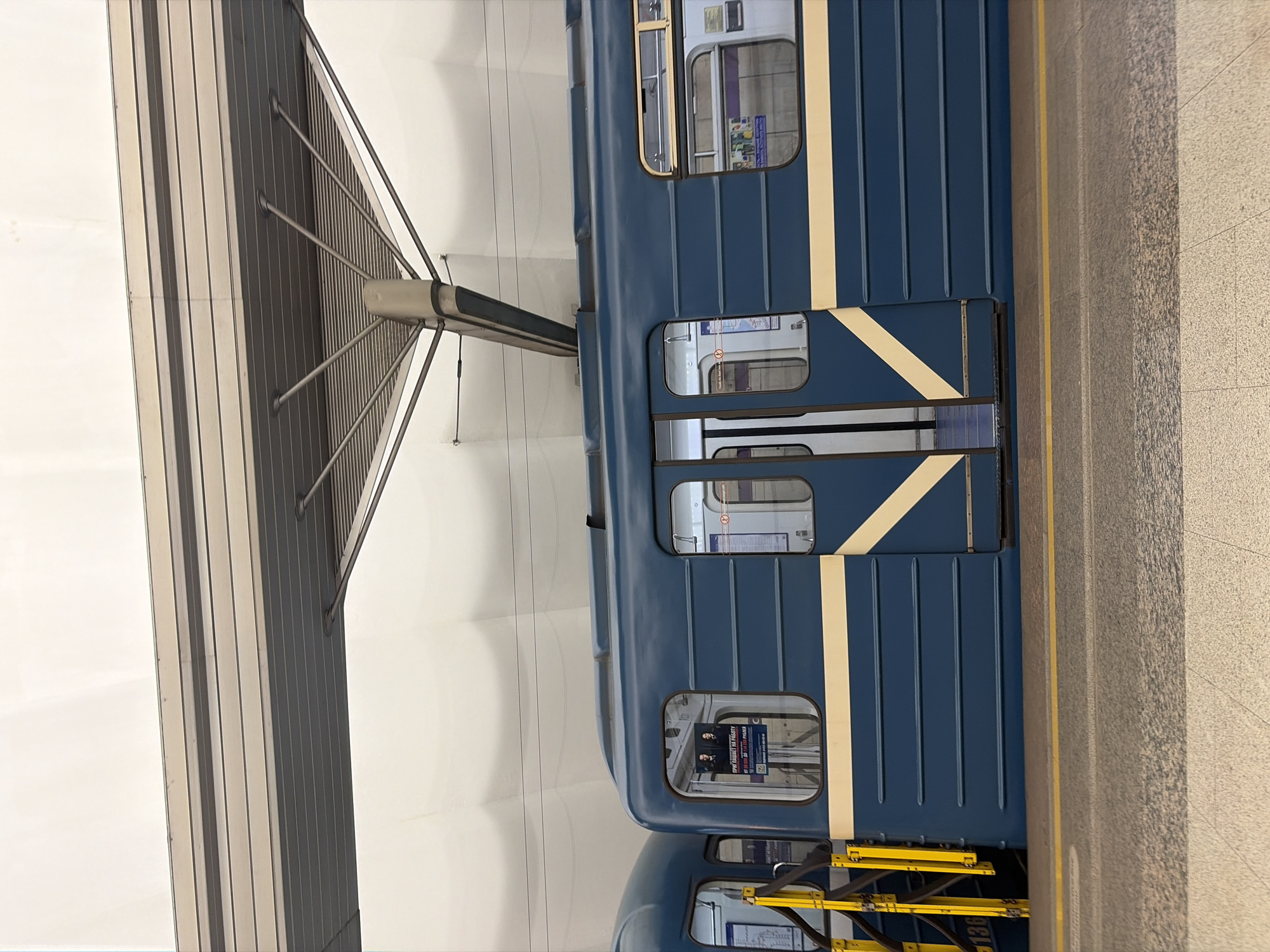 A blue subway train is parked at a station platform, presumably in Shushary, Russia. The train has a light yellow stripe running vertically along the side. Windows show glimpses of the interior and exterior of the train. The platform is paved with grey stone. A metal structure is above the train tracks.