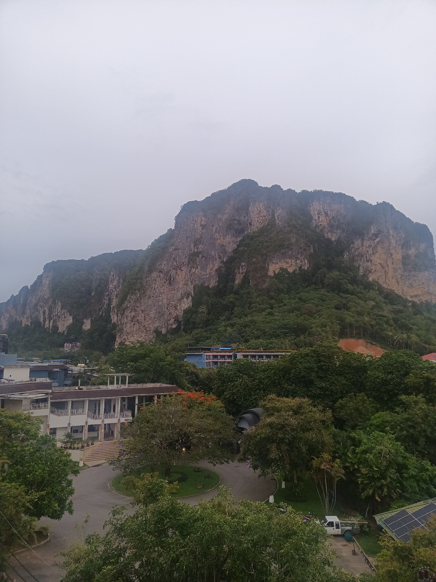 The image shows a view in Ban Ao Nang, Thailand, featuring a large limestone karst mountain as a prominent backdrop. The weather appears overcast. In the foreground, there is a view of buildings and landscaping, including a circular driveway with a grassy island in the center. The buildings appear to be a hotel or resort, with a white facade and columns. Dense green trees and foliage fill the space between the buildings and the mountain, obscuring some of the structures. In the lower right corner, there are solar panels. A white truck is parked near the panels. There are no visible people in the scene.