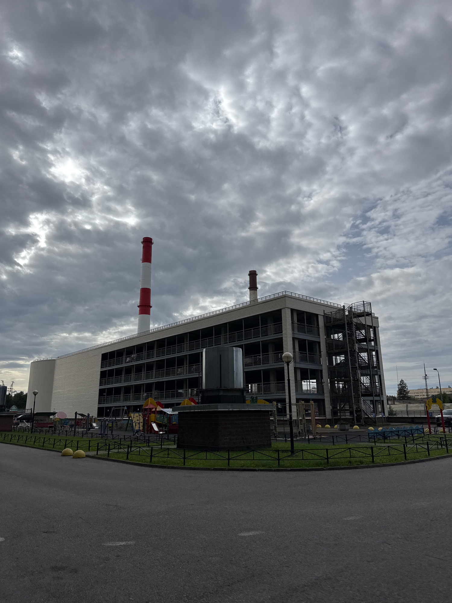 The image depicts a multi-story building with a red and white striped smokestack prominently visible. The building appears to be an industrial or utility structure, possibly a power station or factory, given the smokestack. It has multiple levels with open-air balconies or walkways. An external metal staircase is attached to one side of the building. In front of the building, there is a playground area with swings, slides, and climbing structures. The ground in front of the playground is covered with grass and a low black metal fence. A paved road or pathway curves in the foreground. Two yellow spherical bollards are positioned on the grass. The sky is overcast with heavy, grey clouds. The scene is located in Ladozhskoe Ozero, Russia.