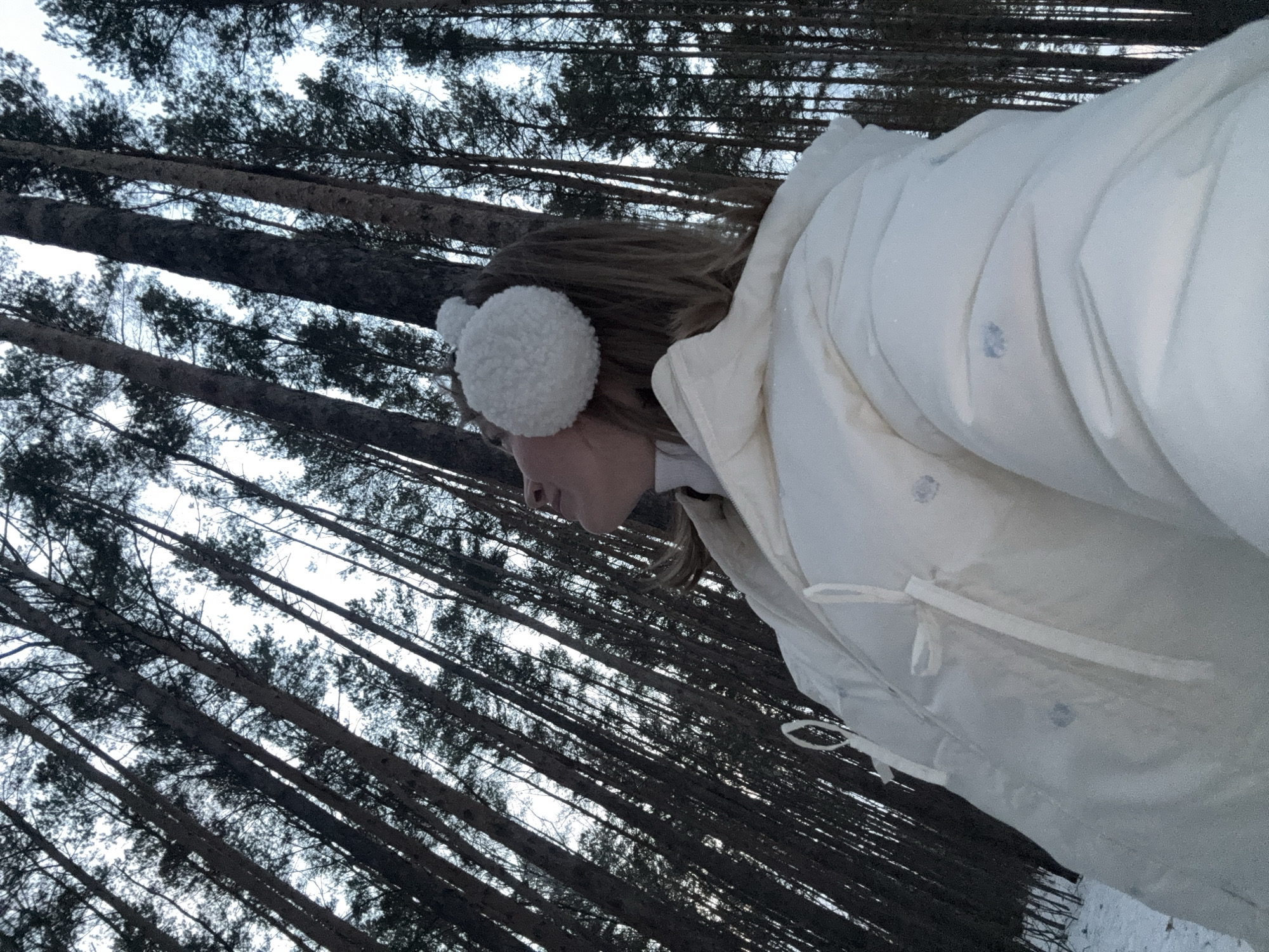 A woman wearing a white coat and white earmuffs is standing in a forest of tall, slender trees. The trees are dark and dense, reaching upwards towards a pale sky. The scene appears to be outdoors, possibly in a wooded area near the city of Chaikovsky, Russia.