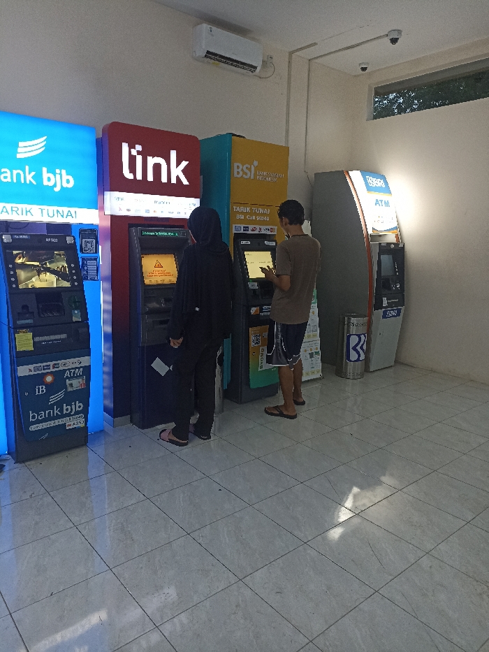 The scene shows three automated teller machines (ATMs) in an indoor location. The location is in Jatinangor, Indonesia, at the coordinates -6.931 latitude and 107.776 longitude. Two people are present. A woman is standing in front of a "tlink" ATM, facing the machine and interacting with it. A man is standing in front of a "BSI" ATM, using it. The "tlink" ATM is to the left of the "BSI" ATM. A third ATM, labeled "ATM" is situated on the right side of the image. The floor is tiled and there is a white wall behind the ATMs. There is a small trash can with a blue recycle symbol on it, situated near the "ATM" machine. The lighting is fluorescent, with some natural light coming in from the left. The atmosphere is calm and quiet.