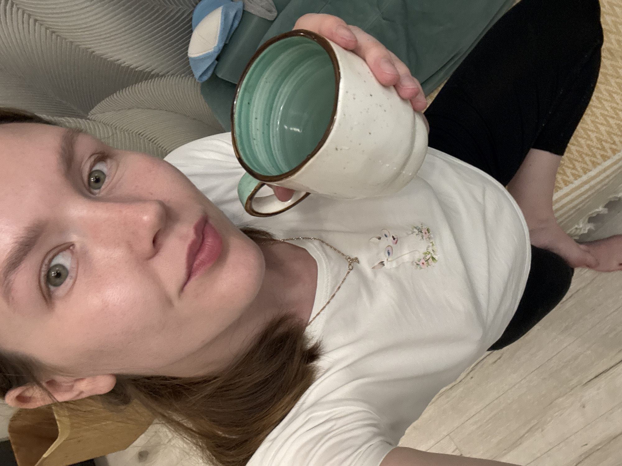 A young woman with light brown hair tied back in a ponytail is holding a white and green speckled mug in her right hand. She is wearing a white t-shirt with a pendant that appears to be a cat with glasses and flowers. She is seated on a floor with light-colored wooden planks. To her right, there is a teal-colored sofa and a blue and white hat. In the background, to the left, a woven basket is partially visible. The lighting suggests an indoor environment, possibly a living room. The scene is set in Izhevsk, Russia.