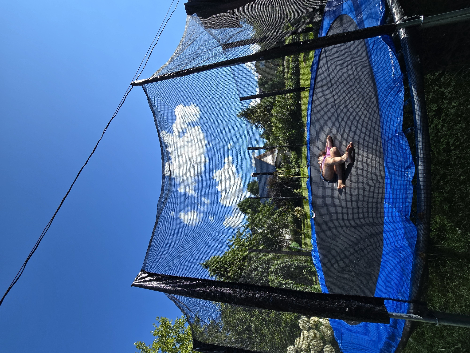 A young person is lying on their stomach on a trampoline in a backyard. They are wearing a pink bathing suit and appear to be resting. The trampoline has a black jumping mat and blue padding around the edge. A safety net encloses the trampoline. The scene is set on a sunny day with a clear blue sky and fluffy white clouds. Green trees and some houses are visible in the background, suggesting a suburban or rural location. The overall mood is peaceful and relaxed, indicating a leisurely summer day. The image is oriented sideways.