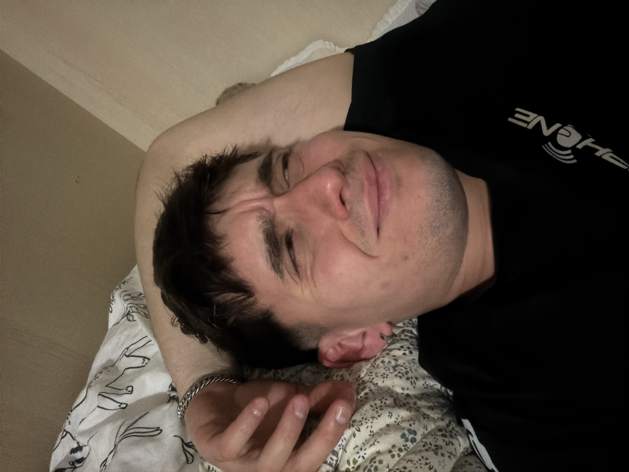 A man is lying on a bed, resting with his arm positioned above his head. He is wearing a black t-shirt and has a silver bracelet on his wrist. The bed is covered with a patterned sheet. The room has a beige wall. There is no information to suggest this is in Sofia, Bulgaria.