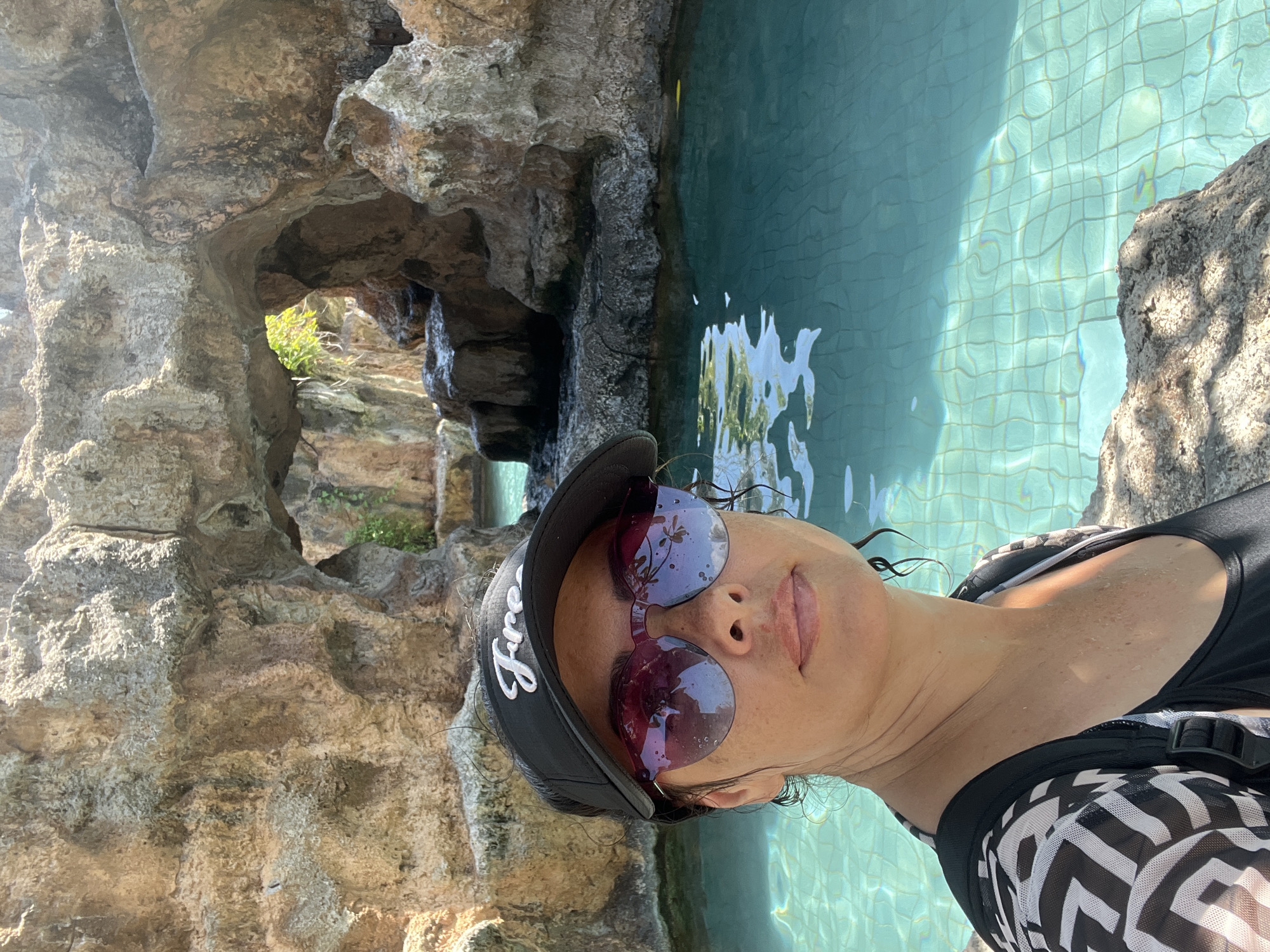 A woman with dark hair and sunglasses is taking a selfie in a swimming pool. She is wearing a black visor and a black and white patterned cover-up over a black swimsuit. The pool is turquoise and appears to be in a tropical setting with rock formations and greenery in the background. The location is the Ayana Resort and Spa in Bali, Indonesia.