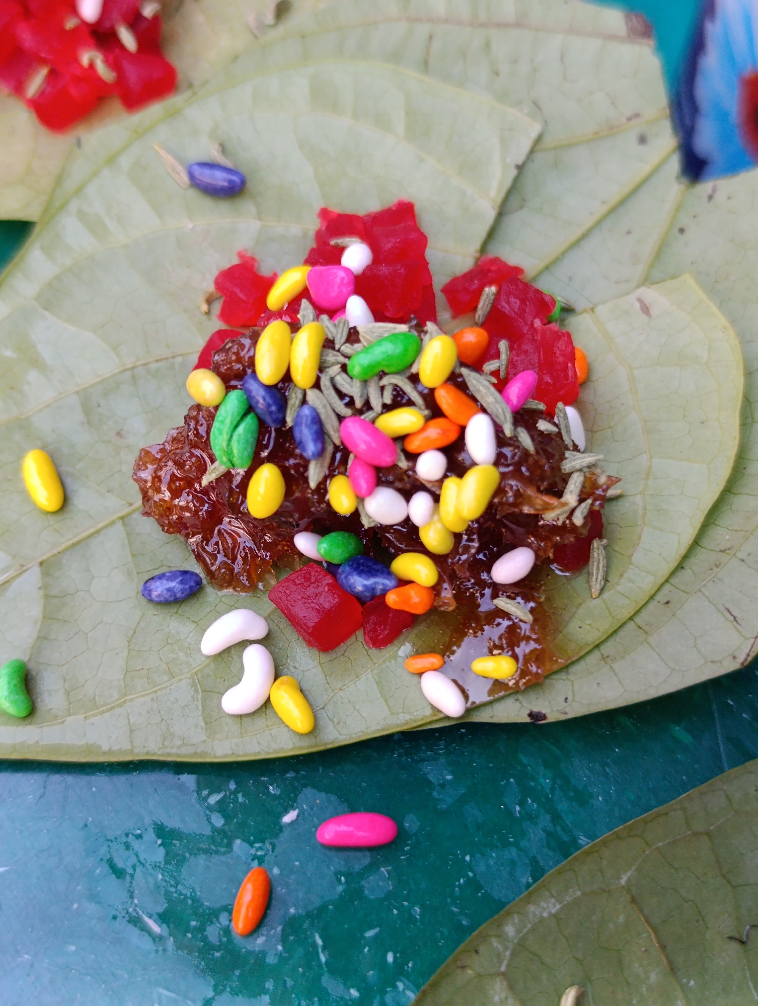 A close-up, overhead view displays an arrangement of sweet or paan ingredients on several light green leaves. A central mound of glossy, dark reddish-brown, viscous material, resembling gulkand, is prominently featured. Adorning this mound and scattered on the surrounding leaves are various components: numerous small, sugar-coated candies in oval and kidney-bean shapes, presenting a vibrant palette of yellow, pink, orange, green, blue, purple, and white. Interspersed among these are bright red, translucent, irregularly shaped or cubed pieces of candied fruit. Elongated, pale green to light brown fennel seeds are also distributed across the central mixture and leaves. Additional individual candies of varying colors (pink, orange, yellow, white, blue, green) are strewn on the underlying dark green, subtly reflective surface. Blurry red elements are partially visible in the upper left background, and a fragmented, indistinct blue and red object appears in the upper right. No individuals are depicted, and no specific actions are occurring.