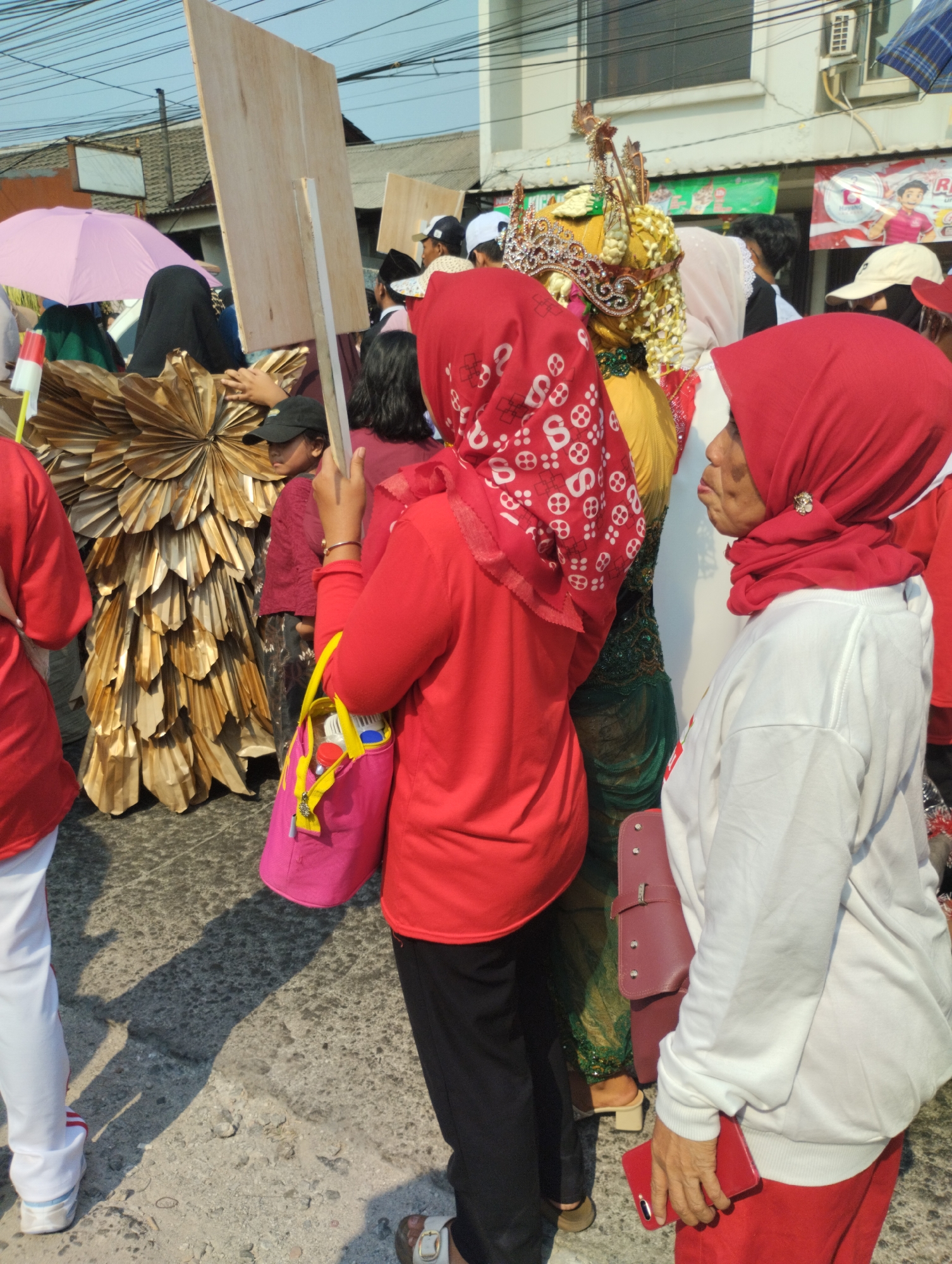 In Bekasi, Indonesia, a group of people are gathered outdoors, possibly for a parade or event. A person in the foreground wears a red headscarf and a white sweatshirt, holding a red smartphone. Beside them, another person in a red long-sleeved shirt and black pants carries a bright pink bag. Behind them, a person in a traditional ornate golden headdress with intricate beadwork stands out. To the left, a pink umbrella is visible, and a large, fan-like golden costume is also present. The ground appears to be paved with rough stones, and overhead, electrical wires crisscross the sky. Buildings are visible in the background.
