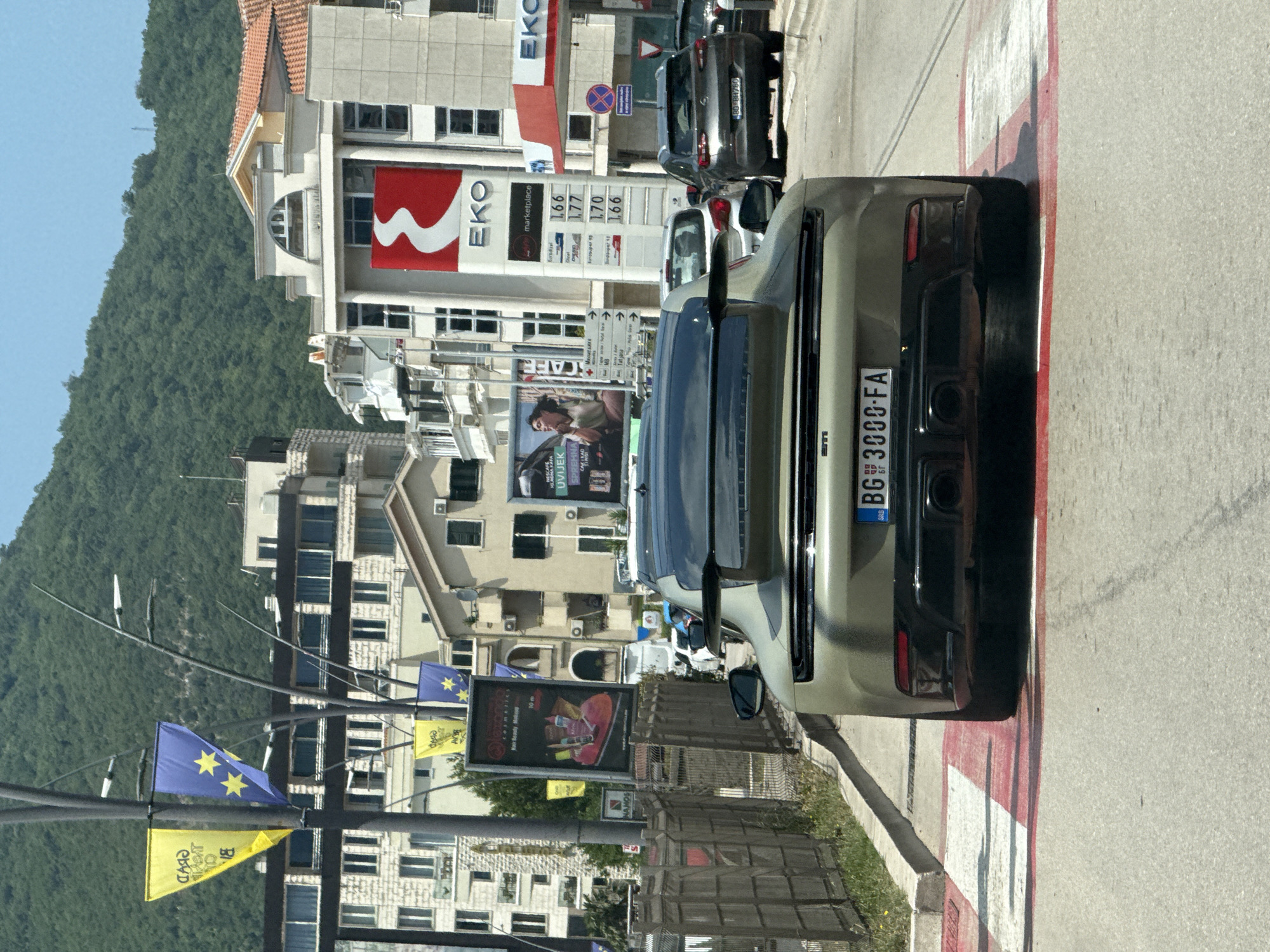 A rear-view shot captures a modern, olive-green Porsche 911 parked on a pedestrian crossing in the city of Budva, Montenegro. The car's license plate reads "BG 3000 FA". Behind the Porsche, other vehicles are visible on the road, including a dark grey SUV with license plate "BG BH786" in the right lane.
The background is a bustling urban scene dominated by multi-story buildings. A prominent EKO gas station sign, featuring a distinctive red and white logo, stands tall among the structures. Flags are mounted on lampposts, with yellow and blue banners adorned with stars. Green, tree-covered hills form the backdrop against a clear blue sky. The weather appears to be sunny. The street is paved, and a metal fence runs along the left side of the road. The image was taken during daylight hours.