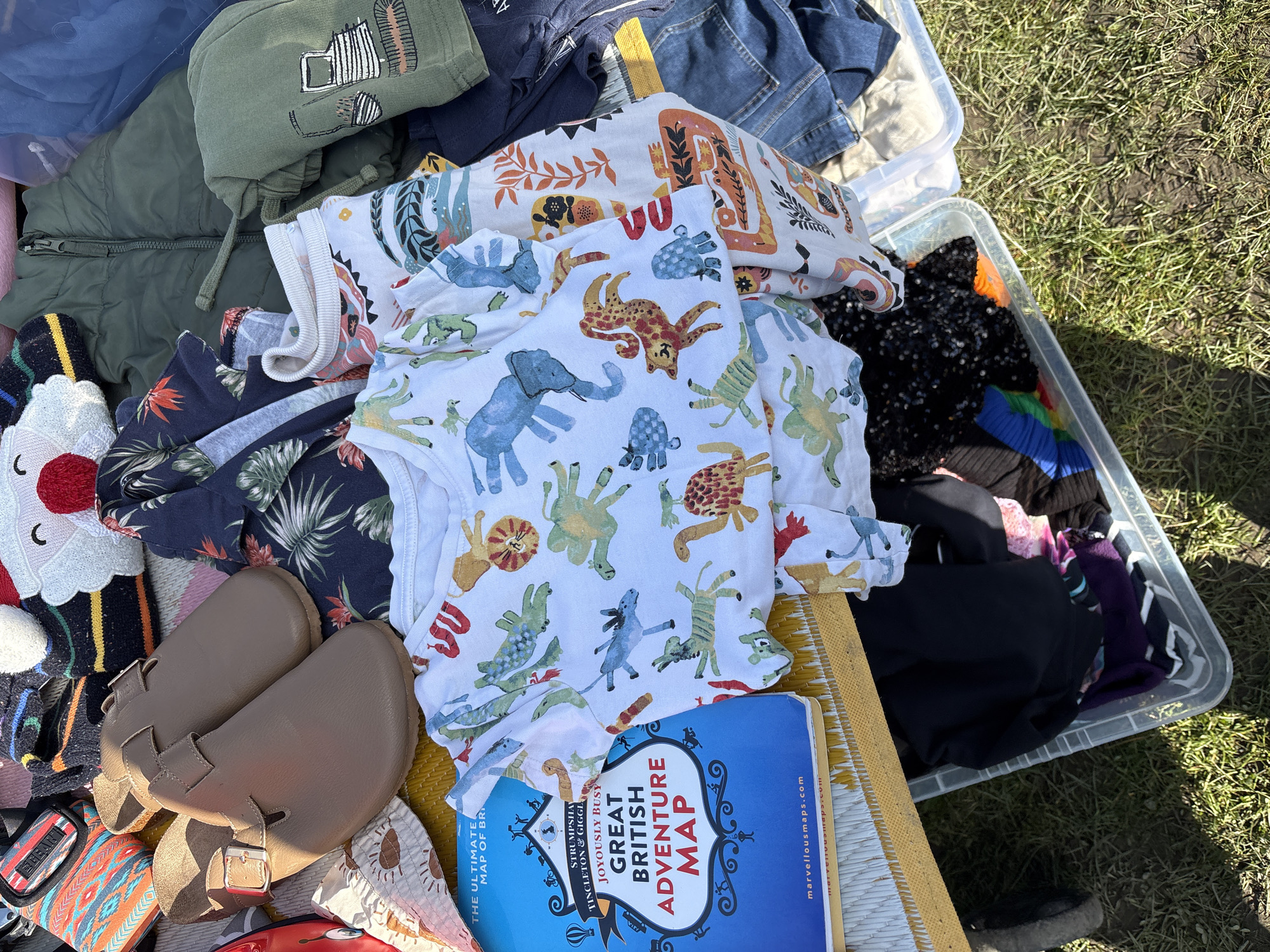 The image captures an outdoor scene with an assortment of items placed on a patterned mat or table, suggesting a sale or display. The scene includes various clothes, with a white onesie featuring animal patterns prominently positioned. Among the clothes, there is a stack of tops, a green jacket, and other colorful garments. A pair of brown leather-like shoes sits on the mat, adding to the collection of items.
A blue map book titled "Great British Adventure Map" is partially visible at the bottom, indicating the location is in the United Kingdom. Based on the presence of the "Great British Adventure Map," it is probable that the items are being displayed or sold in a town or city within the UK. Although Chirk is a town in the United Kingdom, there is no definitive information to confirm that this scene takes place specifically in the city of Chirk. The background shows a glimpse of grassy terrain, suggesting an outdoor setting. The overall scene gives the impression of a collection of items laid out for sale or organization.