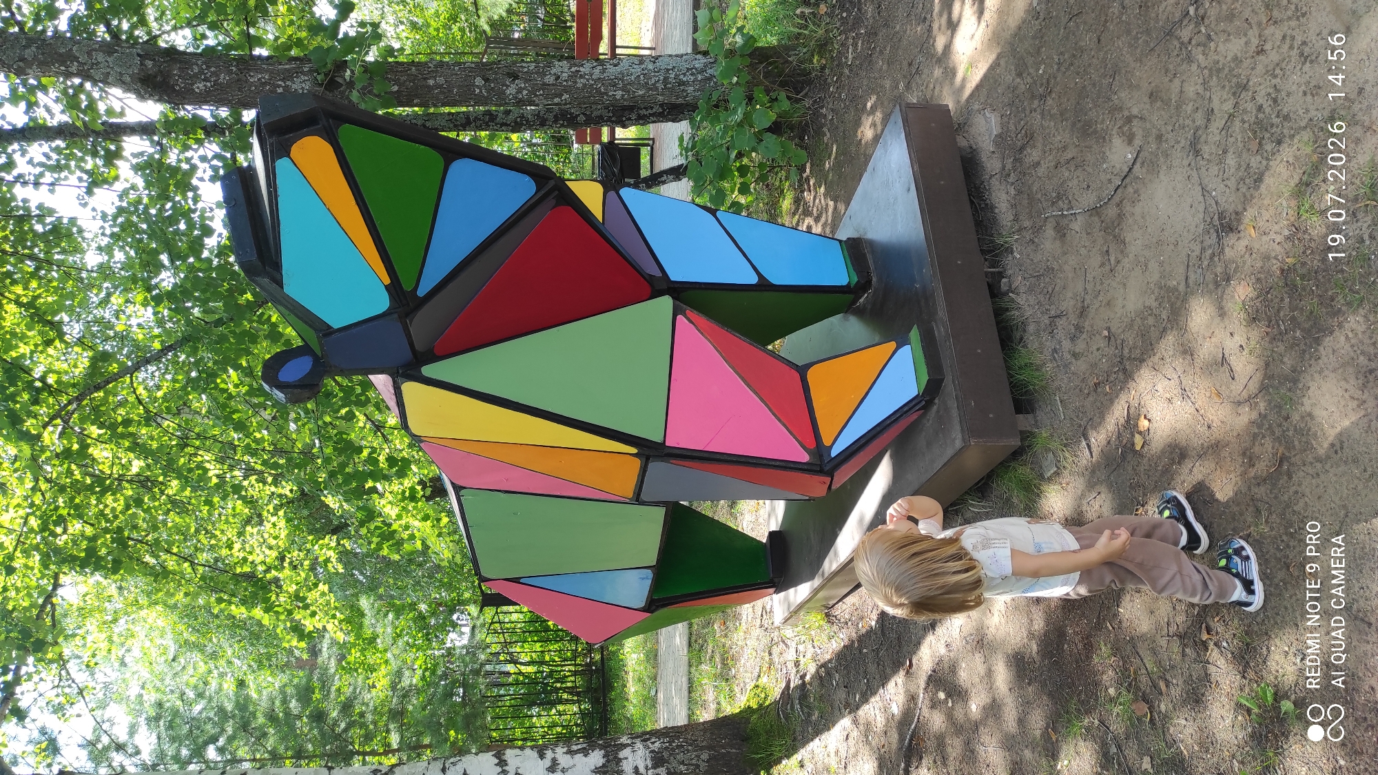 A young child with blonde hair, wearing a light-colored t-shirt and brown pants, stands in a park looking at a large, geometric sculpture of a bear. The sculpture is composed of many colorful, faceted shapes in red, blue, green, yellow, pink, and orange. The bear sculpture is mounted on a dark base. The child is wearing dark sneakers with green accents. The park setting includes trees with green foliage, a wooden fence, and a wooden bench in the background. The ground is covered in dirt and scattered leaves. The timestamp in the lower right corner indicates the photo was taken on July 19, 2026, at 14:56. The watermark in the lower left corner indicates the photo was taken with a Redmi Note 9 Pro. The location is identified as the city of Chaikovsky, Russia.