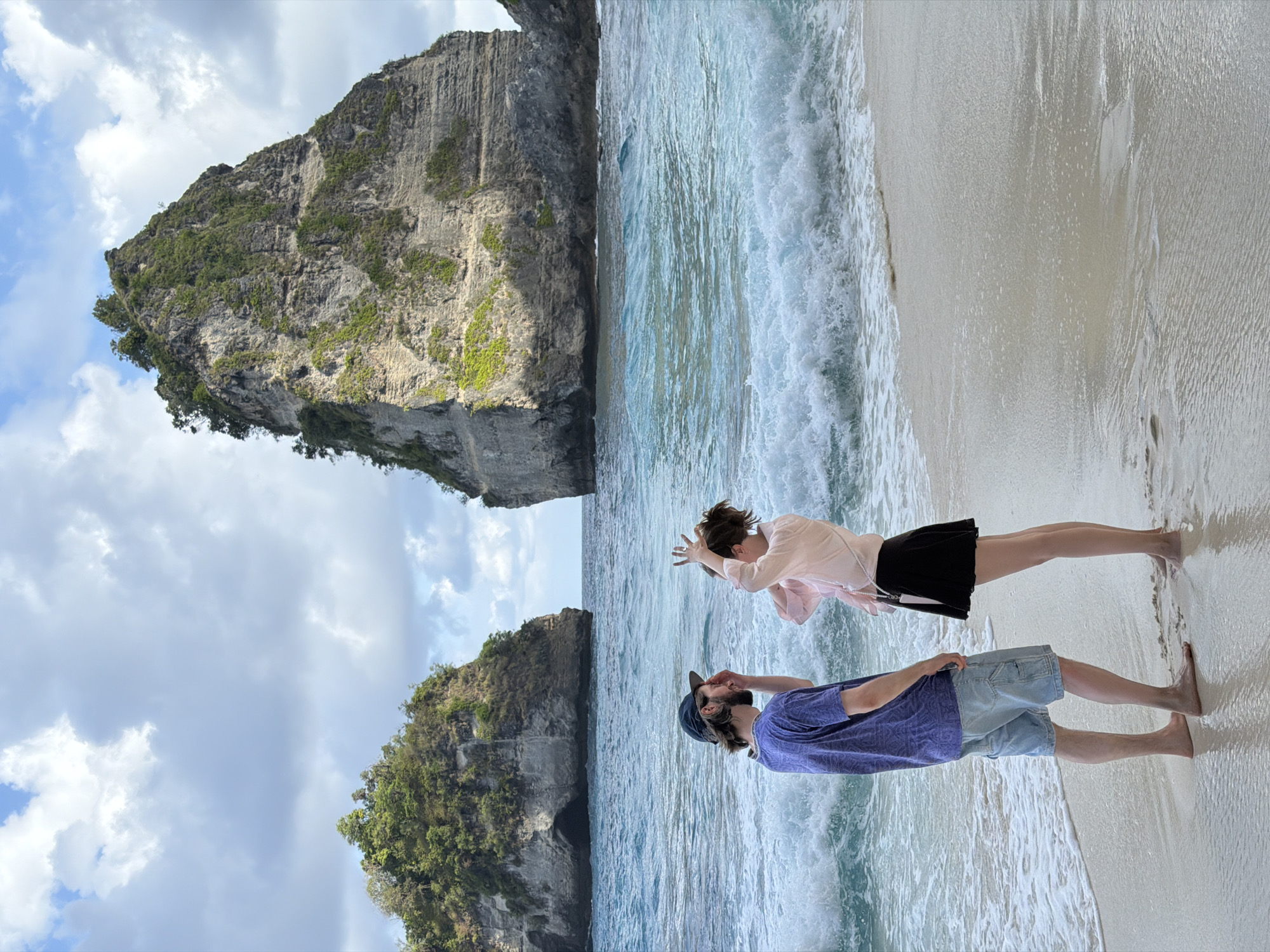 The image shows two people standing on a sandy beach with waves gently lapping at the shore. In the background, two large, lush green rock formations rise dramatically from the turquoise ocean. The sky is partly cloudy with patches of blue visible. The person on the left, a male, is wearing a blue t-shirt and denim shorts and a blue baseball cap. He is facing towards the right, with his right hand near his face. The person on the right, a female, is wearing a light pink shirt and a black skirt. She is facing the male and has her hands raised near her head, as if posing or making a gesture. The overall scene depicts a serene beach environment, likely in Nusa Penida, Indonesia, a location known for its stunning natural rock formations and beautiful coastlines.