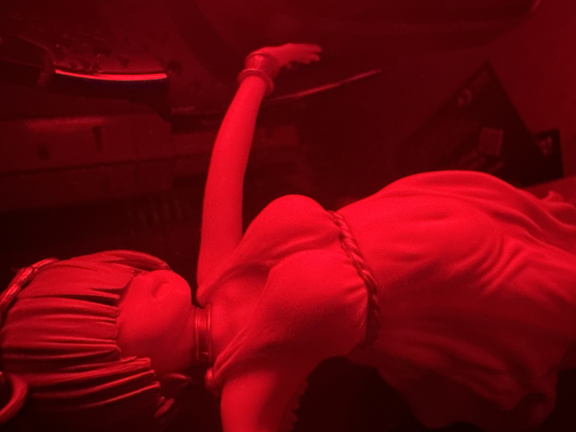The image is a close-up, dimly lit, and vertically oriented photograph of a figurine. The entire scene is bathed in a strong red light, which obscures fine details and creates a monochromatic effect. The figurine appears to be of a female character with a stylized hairstyle and a draped garment. The garment has noticeable folds and appears to be cinched at the waist by a rope-like belt. The figurine is positioned as if in motion, with one arm extended to the right, adorned with a bracelet. The background is dark and out of focus, offering no discernible details of its surroundings. The location is Unknown, Unknown.