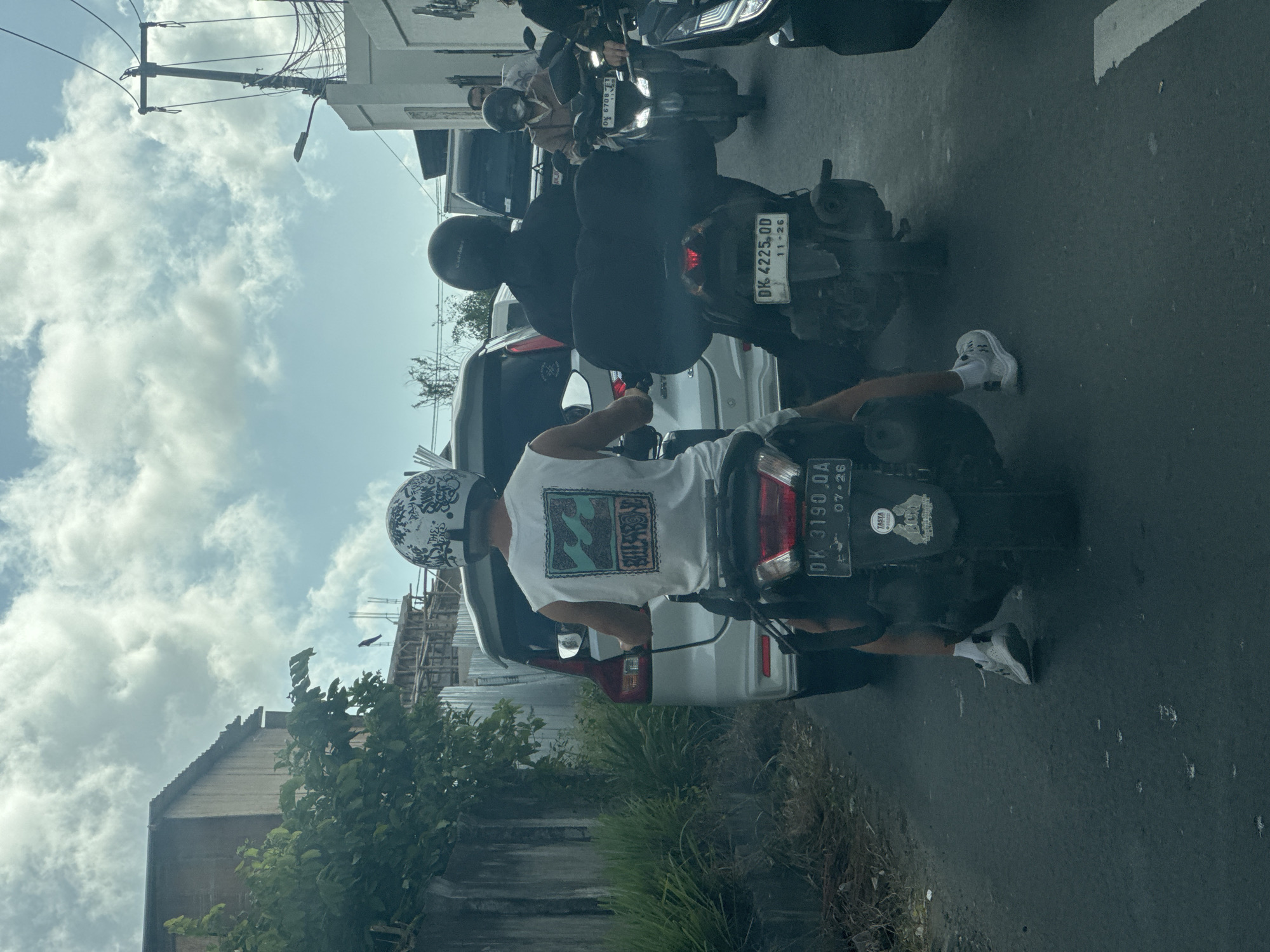 The image depicts a street scene in Kuta Selatan, Indonesia, with several motorbikes and a white vehicle amidst traffic. Two motorbikes are prominently positioned in the foreground. One motorbike has a license plate reading "DK 3190 OA 07•26," and the rider is wearing a white tank top with a graphic print and a helmet with a black and white design. Another motorbike directly ahead has a license plate reading "DK 4225 OD 11•26," with its rider wearing a black helmet and a black cloth covering an oversized black bag on the seat. The background reveals a partially obscured white car, a building under construction, and a line of utility poles against a partly cloudy sky. Vegetation is also visible along the roadside.