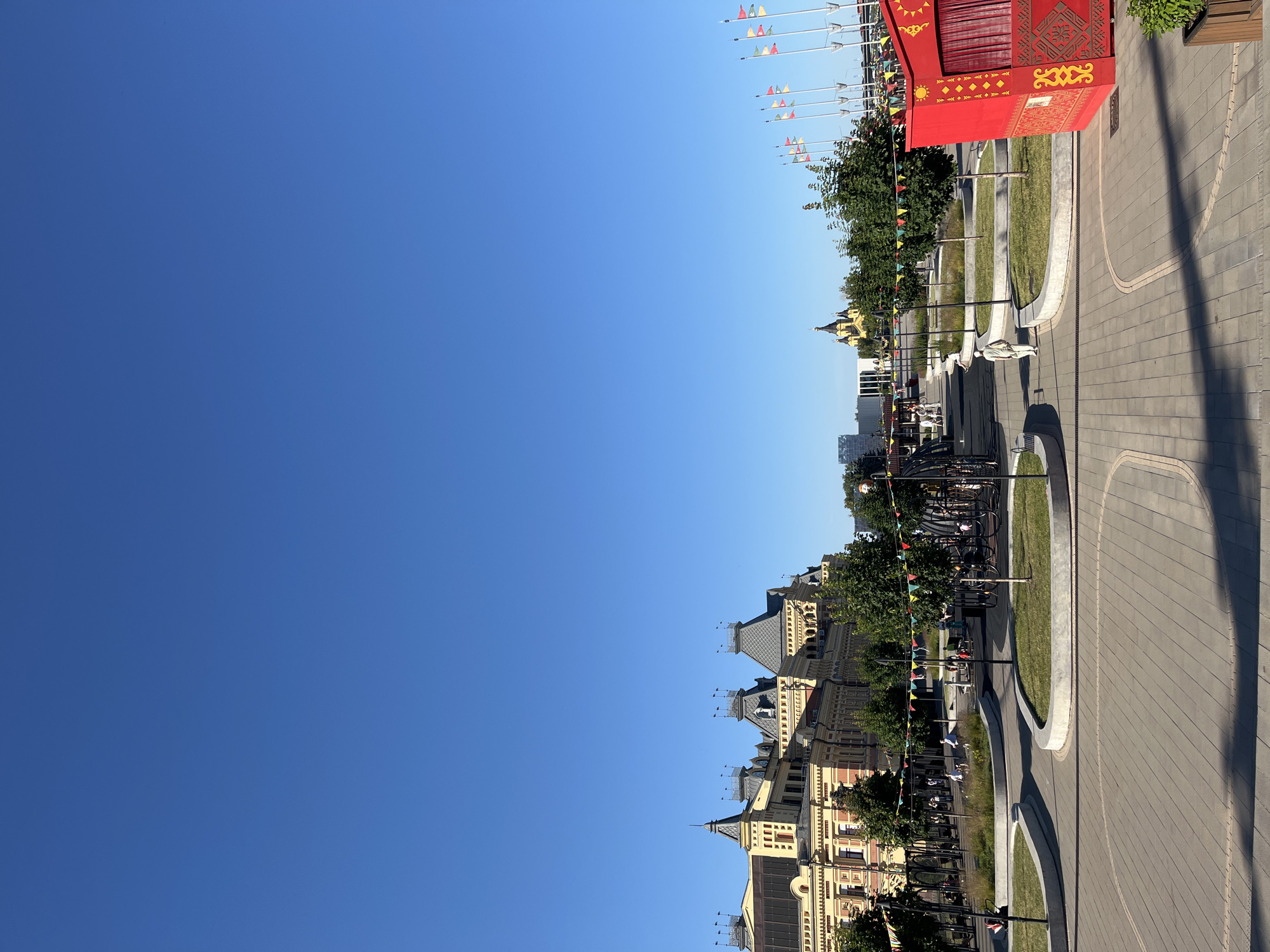 This is a low-angle, landscape-oriented photograph taken on a clear, sunny day, likely in the late morning or early afternoon given the bright light and distinct shadows. The scene depicts an outdoor public space in Nizhny Novgorod, Russia, featuring a blend of modern urban design and traditional architecture.
On the right side of the image, a paved walkway with a distinct curvilinear pattern leads into the distance. Adjacent to the walkway, a manicured lawn area is bordered by curved concrete structures. A bright red, ornate structure, possibly a kiosk or a temporary stage, is positioned prominently in the foreground, adorned with yellow decorative patterns and fabric curtains. Flags on poles line the edge of the walkway, adding a festive atmosphere.
In the background, a row of classic European-style buildings with mansard roofs and light-colored facades stretches across the frame. These buildings suggest a historical district or a significant architectural area within the city. Green trees provide a natural element, interspersed between the buildings and along the walkway.
Several people are visible in the scene, appearing as small figures engaged in leisurely activities. Some are walking along the pathway, while others are seated on benches or interacting in small groups, conveying a relaxed and pleasant mood. The overall impression is of a well-maintained public square or park designed for recreation and enjoyment, likely a popular spot for both locals and visitors. The clear blue sky indicates fair weather.