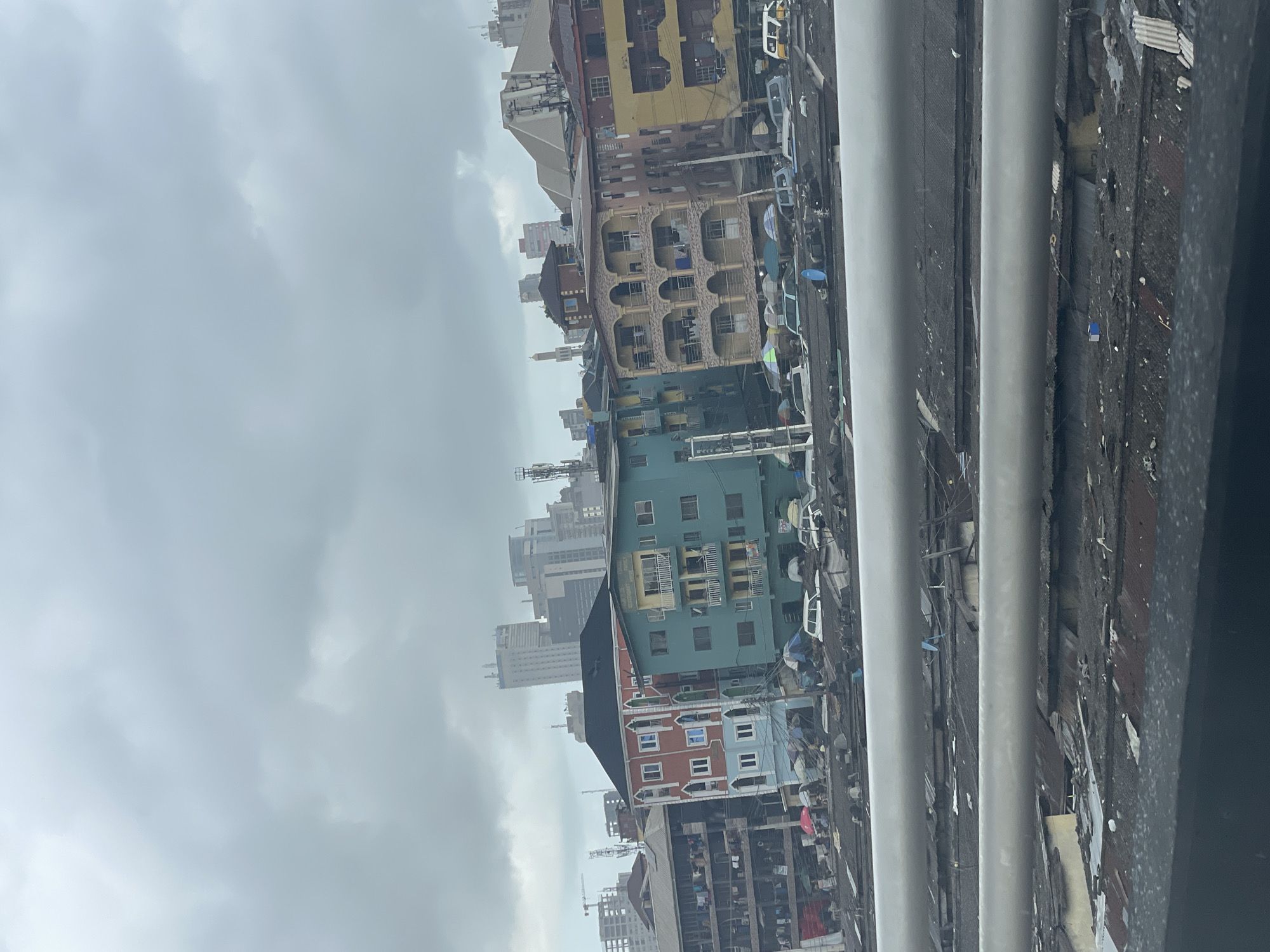 The image presents a view of Lagos, Nigeria, under a cloudy sky, as observed from an elevated position. The foreground shows elements of the structure from which the photo is taken, featuring two vertical pipes and decaying corrugated iron sheets. Beyond this, a congested urban landscape unfolds, characterized by a mix of medium-rise buildings in various states of repair, painted in colors such as yellow, blue, and orange. Evidence of dense inhabitation is visible, including clotheslines with laundry hanging. Interspersed among the buildings are parked vehicles, their types obscured by the distance and the overcast conditions. In the background, taller buildings can be discerned, indicating the presence of a central business district or more modern developments further away. The overall impression is one of high-density urban living, set against a backdrop of a grey and overcast Lagos sky.
