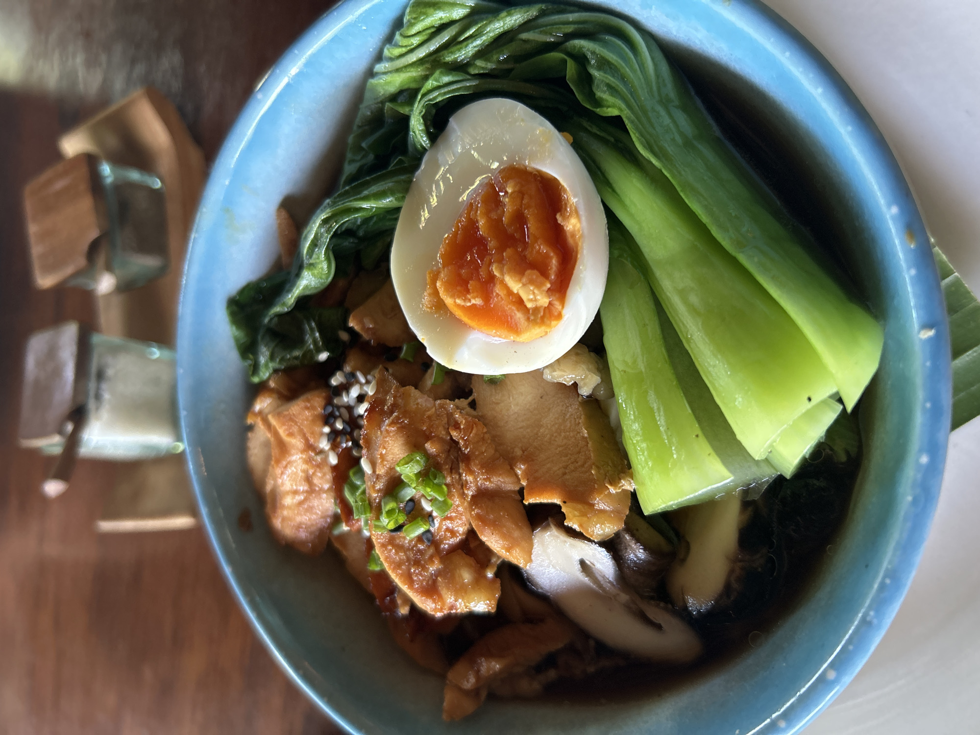 A close-up, eye-level, indoor shot shows a bright blue ceramic bowl filled with a savory Asian dish. The bowl is positioned on a wooden table, with blurred salt and pepper shakers in glass containers with wooden lids in the background, suggesting a restaurant setting. The dish features sliced, glistening pieces of what appears to be chicken or pork, intermingled with shiitake mushrooms and dark leafy greens, possibly bok choy. A perfectly halved hard-boiled egg with a vibrant orange yolk sits prominently on the right side of the bowl. The dish is garnished with chopped green onions and black and white sesame seeds. The lighting is natural, creating soft shadows and highlighting the textures of the food. The overall impression is one of a nourishing and appetizing meal.