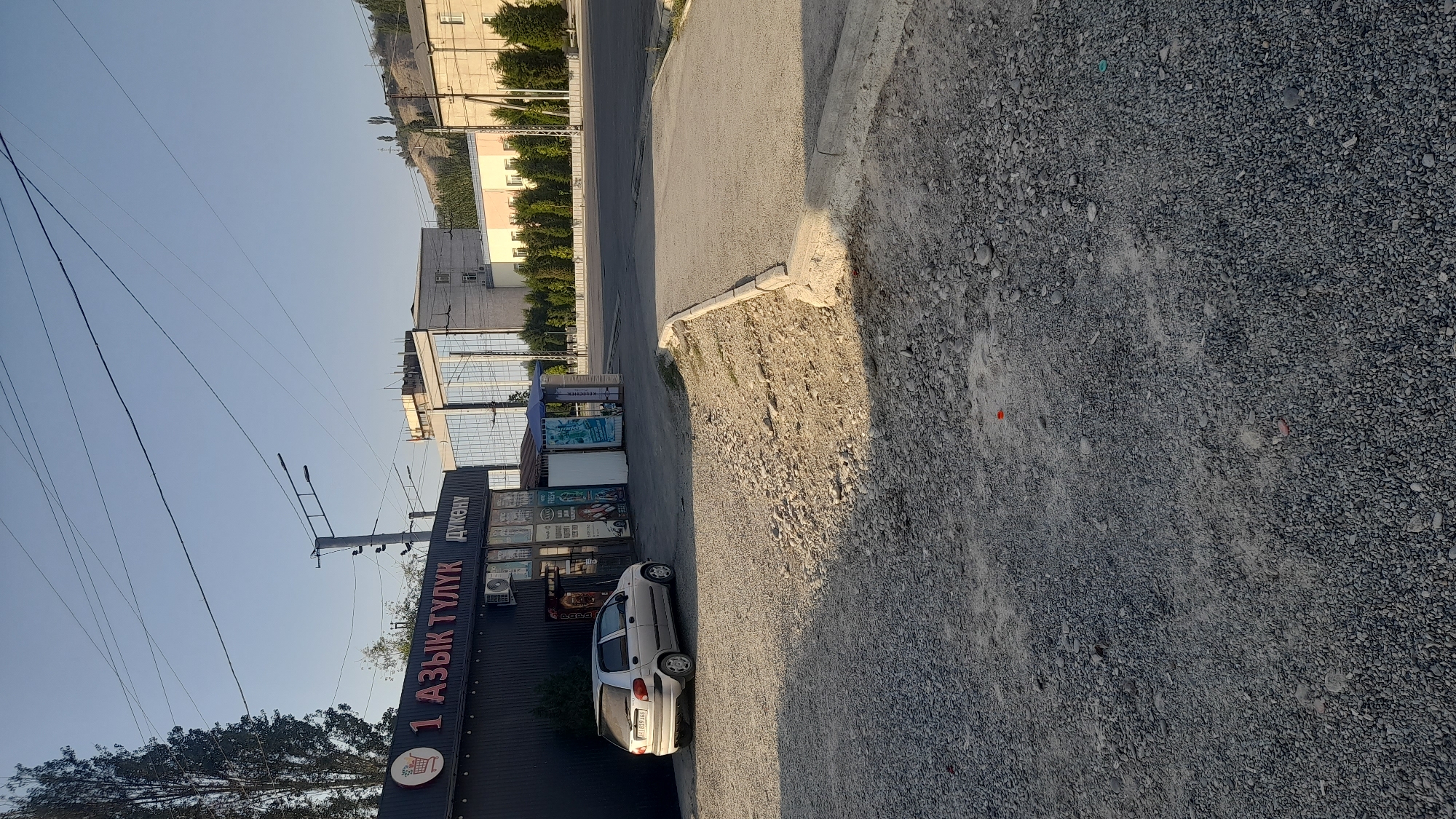 An exterior shot captures a street-level view of a storefront in Osh City, Kyrgyzstan. The storefront, labeled with Cyrillic text "1 АЗЫК ТУЛУК ДҮКӨНҮ", is a dark, rectangular building with large glass windows displaying various products. A silver sedan is parked to the left of the storefront. The ground in front of the store is covered in gravel and dirt, with a concrete curb separating it from the paved road. In the background, several buildings of varying architectural styles can be seen, along with trees and a distant hillside. The sky is clear and blue, suggesting a sunny day. The overall scene depicts a commercial area in a city.