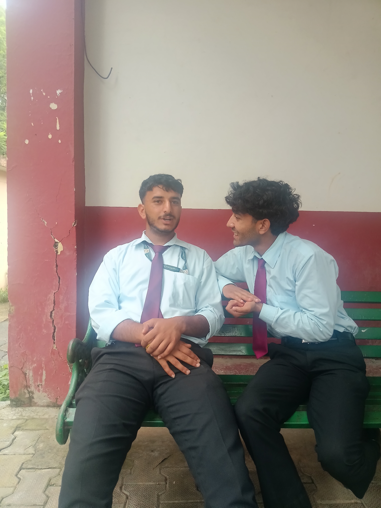 Two young men, dressed in light blue shirts and dark ties, are sitting on a green park bench outdoors. The man on the left, facing the camera, has short dark hair and a mustache. He is wearing a purple tie and black pants, with his hands clasped in his lap. A gold ring is visible on his right ring finger. The man on the right, in profile, has curly dark hair and is looking to his left, smiling. He is also wearing a purple tie and black pants, with his left arm resting on the back of the bench and his right hand touching the left man's arm. Behind them is a wall with peeling red paint on the left and a beige wall on the right. The ground in front of the bench is paved with interlocking concrete tiles. The presence of a red painted structure, similar to architectural elements found in colonial-era buildings, combined with the tiled pavement and general aesthetic, suggests the location could be within the grounds of a government institution or a historical site in Dharamshala, India, potentially near buildings with this architectural style, such as those in the old city area or educational institutions.
