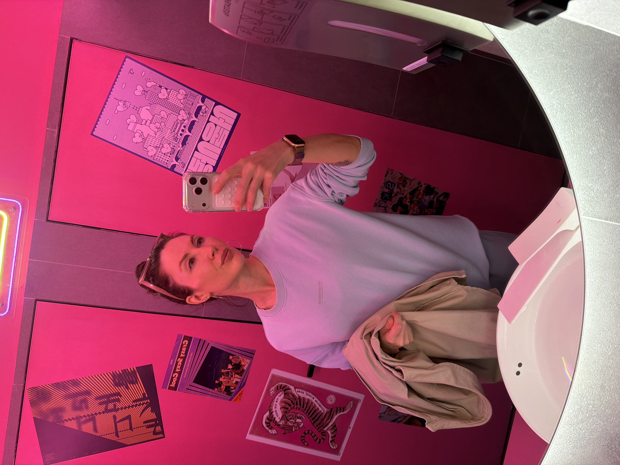 A woman in a powder blue sweatshirt is reflected in a bathroom mirror. She is holding a smartphone in her right hand, apparently taking a selfie. A khaki-colored jacket is draped over her left arm. She is wearing a wristwatch. In the background, the bathroom walls are pink and covered with assorted posters and decorative items. A hand dryer is mounted above the posters. The bathroom is located in Perm, Russia.