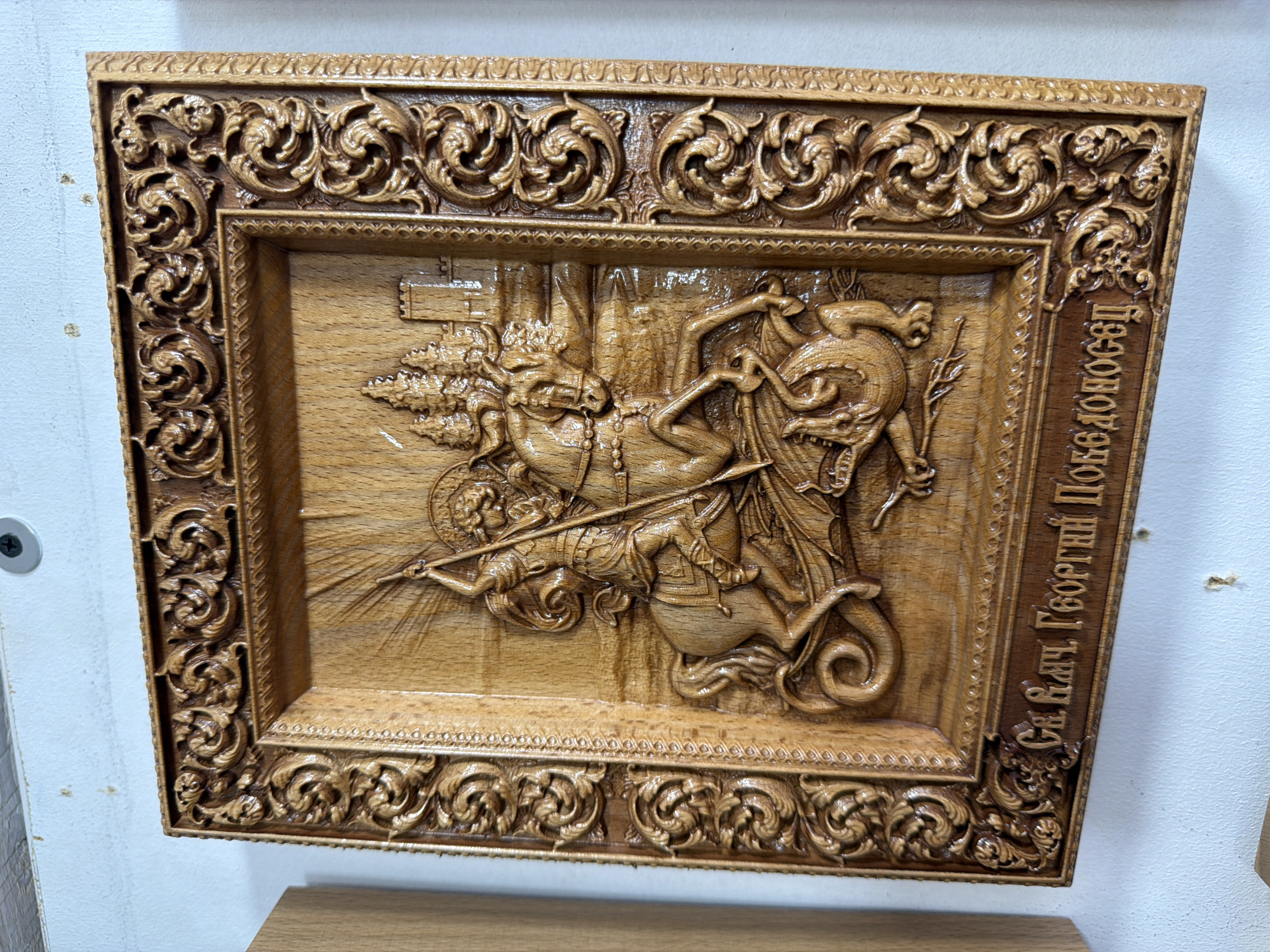 The image shows a carved wooden icon depicting Saint George slaying a dragon. The icon is framed with intricate scrollwork. Below the carving, there is an inscription in Cyrillic script. The icon is mounted on a white wall, with a portion of a wooden object visible to the left. The background appears to be an indoor setting with a white ceiling. No specific location within the city of Yalta, Ukraine can be determined from the image.