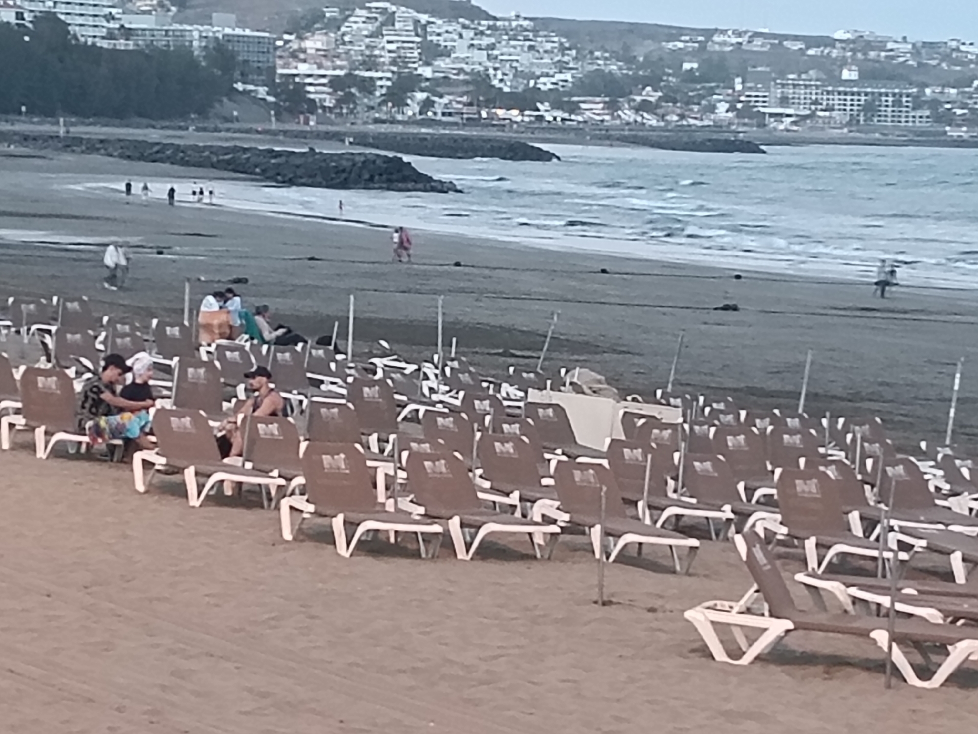 The image shows a beach in San Bartolomé de Tirajana, Spain, filled with rows of empty beach chairs. A few people are sitting in the chairs. In the background, there is a view of the coastline, water, and a cityscape. People are walking along the beach.