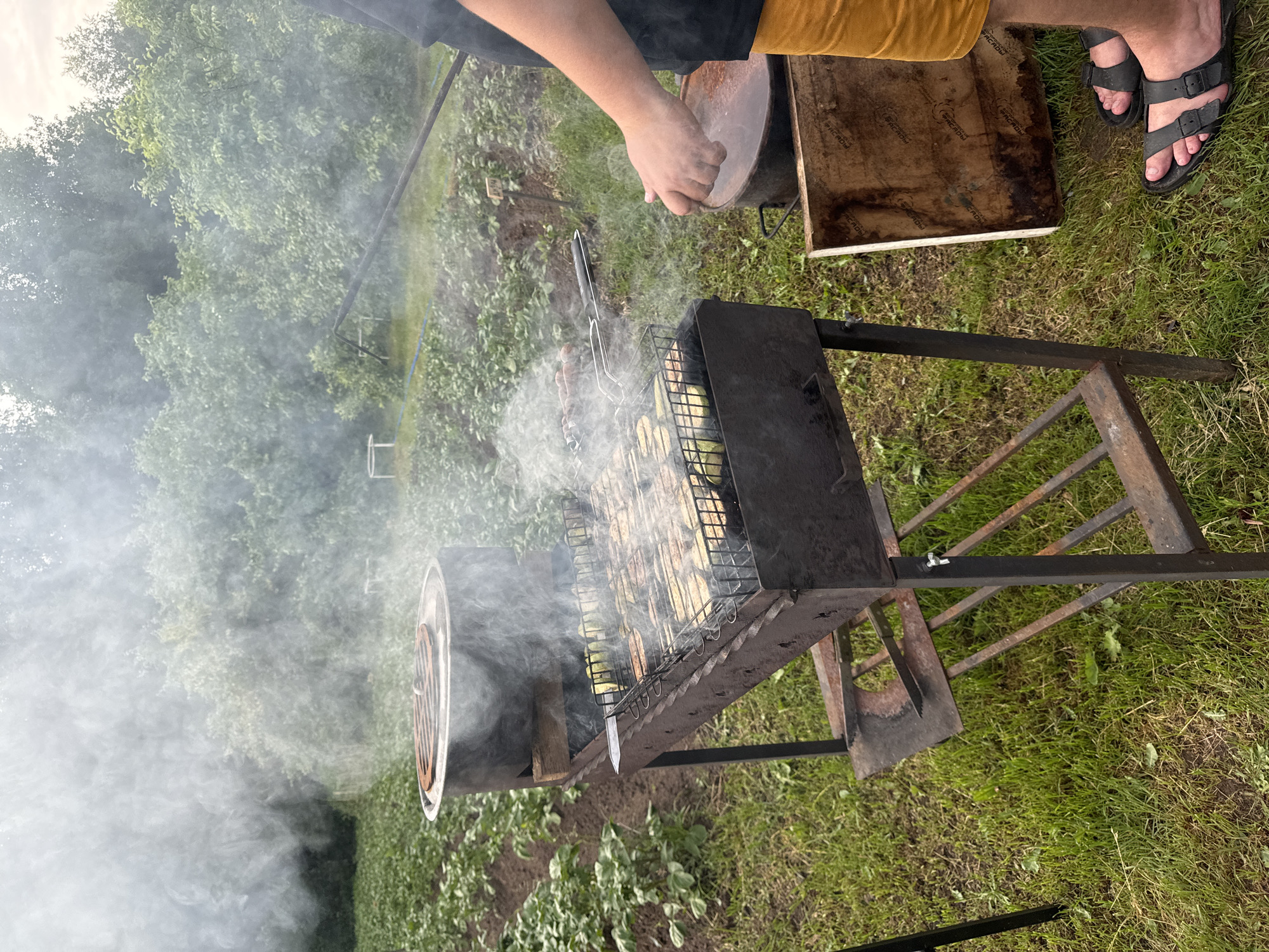 The image depicts an outdoor scene with a charcoal grill in focus. The grill is laden with food, appearing to be vegetables and possibly sausages, cooking over coals and emitting a significant amount of smoke. To the left of the grill, a large cylindrical pot with a lid sits on top of a smaller metal structure. The ground is covered in green grass, and behind the grill, rows of plants are visible, suggesting a garden or agricultural setting. In the foreground, a person's legs are visible, wearing orange shorts and black sandals, with one hand reaching towards the grill. The background consists of lush green trees and foliage, indicating a rural or semi-rural environment. The presence of a grill and garden suggests a domestic or recreational activity, possibly in the village of Ponaryevo, Russia, given the prompt. The overall atmosphere is that of a warm, outdoor cooking event.