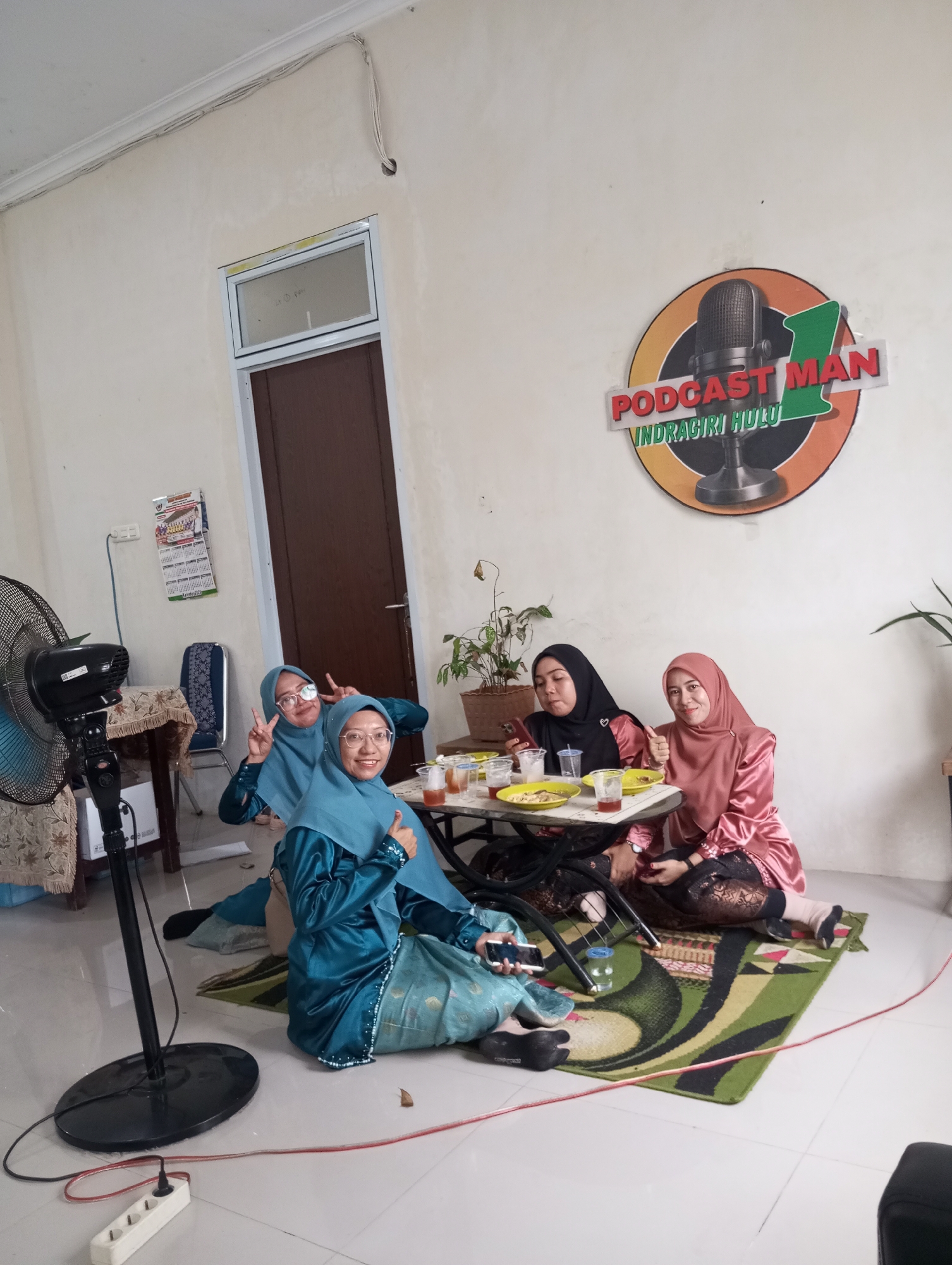 In a room with cream-colored walls and a tiled floor, four young women wearing hijabs are gathered around a small table. The woman on the far left, in a teal hijab and matching outfit, is seated on the floor and holding a smartphone. Next to her, another woman in a teal hijab and outfit is sitting on the floor, making a peace sign with her right hand and a thumbs-up with her left. The third woman, wearing a black hijab and pink satin top, is seated at the table, looking down at her phone. The fourth woman, in a pink hijab and satin outfit, is also seated at the table, giving a thumbs-up with her left hand. A table with plates of food and drinks is in the center of the group. A fan is positioned to the left of the frame, and a power strip with an extension cord runs across the floor. A logo with a microphone and text reading "PODCAST MAN INDRAGIRI HULU" is visible on the wall in the background. The overall scene suggests a casual gathering or meal, possibly related to the podcast mentioned in the logo. This appears to be an indoor setting within a residential or small office space, located in Pematang Reba, Indonesia.