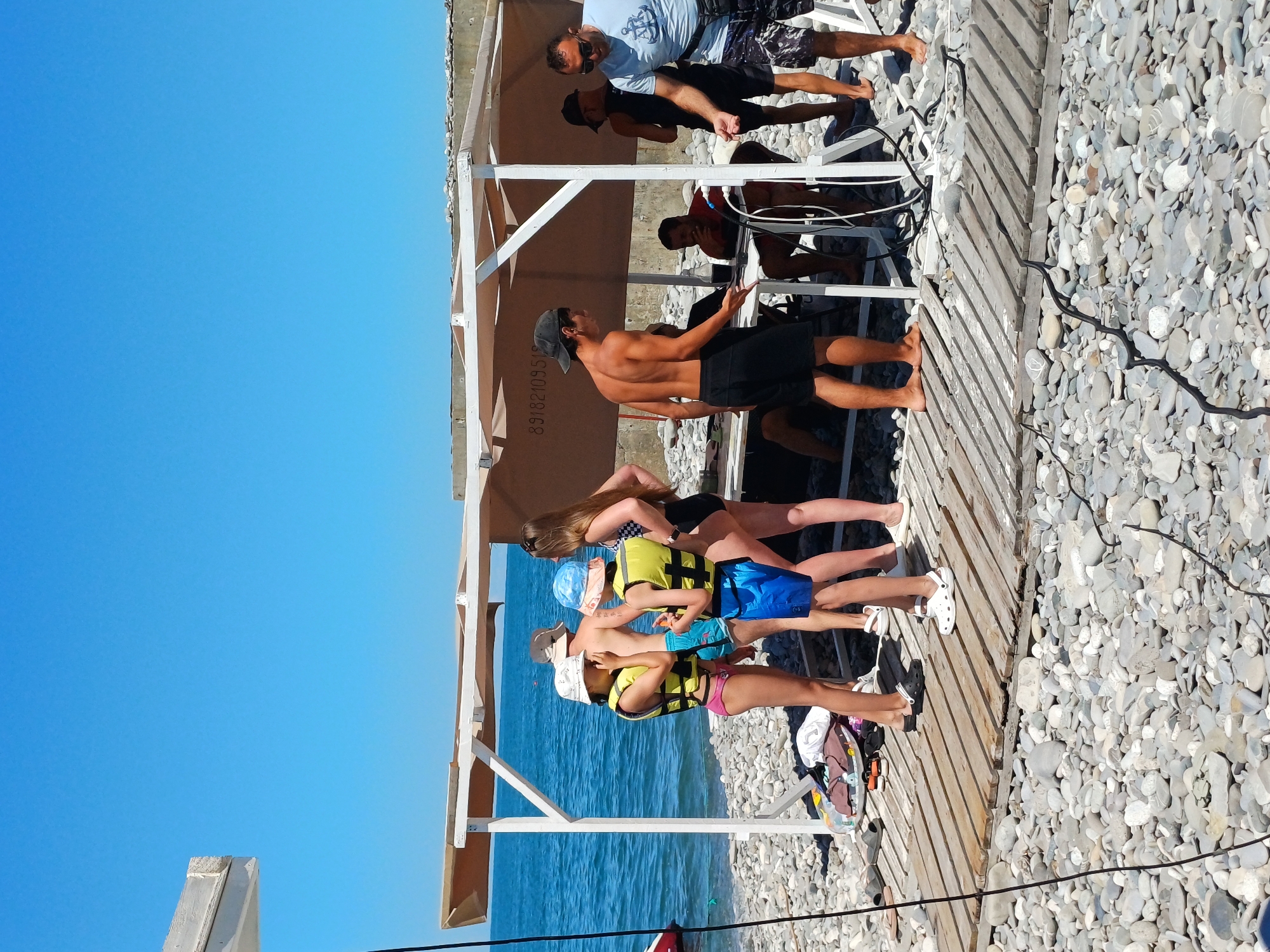 This image depicts a beach scene in Sochi, Russia, on a sunny day. A group of people, appearing to be mostly young adults and children, are gathered near a wooden structure with a canvas roof, likely a beach concession or rental stand.
Several individuals are wearing life vests, suggesting a water sport activity might be taking place or is about to occur. Two children in the foreground are wearing yellow life vests and bucket hats. A young woman in a black and white bikini stands to their right. Further back, a shirtless man in black shorts is engaged in what appears to be a transaction or instruction at a table under the shelter. Other adults are visible in the background, some in swimwear and others in casual clothing.
The environment is a pebble beach, with the turquoise waters of the Black Sea visible behind the people. The ground is covered in smooth, grey and white pebbles. A wooden boardwalk or platform extends from the shelter into the beach area. The sky is a clear, bright blue. Various items such as bags and sandals are scattered on the ground near the shelter. The overall atmosphere suggests a recreational setting on a warm day.