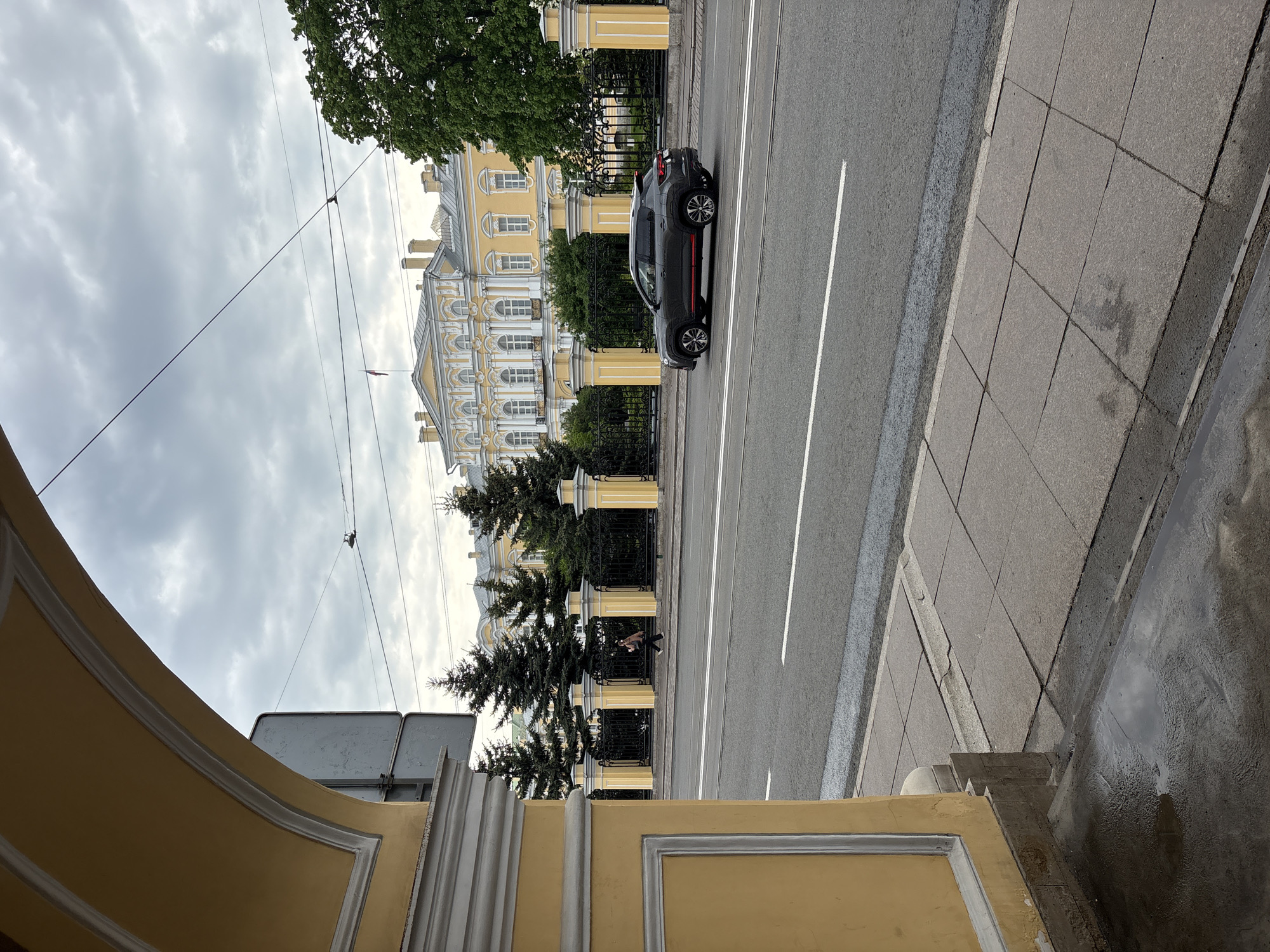 The image depicts a street scene in Saint Petersburg, Russia, likely near the Neva River. A grey car with a red accent stripe drives on an asphalt road with tram tracks and white lane markings. Adjacent to the road is a sidewalk composed of square grey pavers. A low, yellow building with white architectural details frames the foreground.
Across the road, a wrought-iron fence runs along a yellow building with white trim and multiple windows. Behind the fence are evergreen trees. A person can be seen walking behind the fence. Overhead, electrical wires crisscross the sky, which is overcast. The overall environment suggests a blend of historical architecture and urban infrastructure.