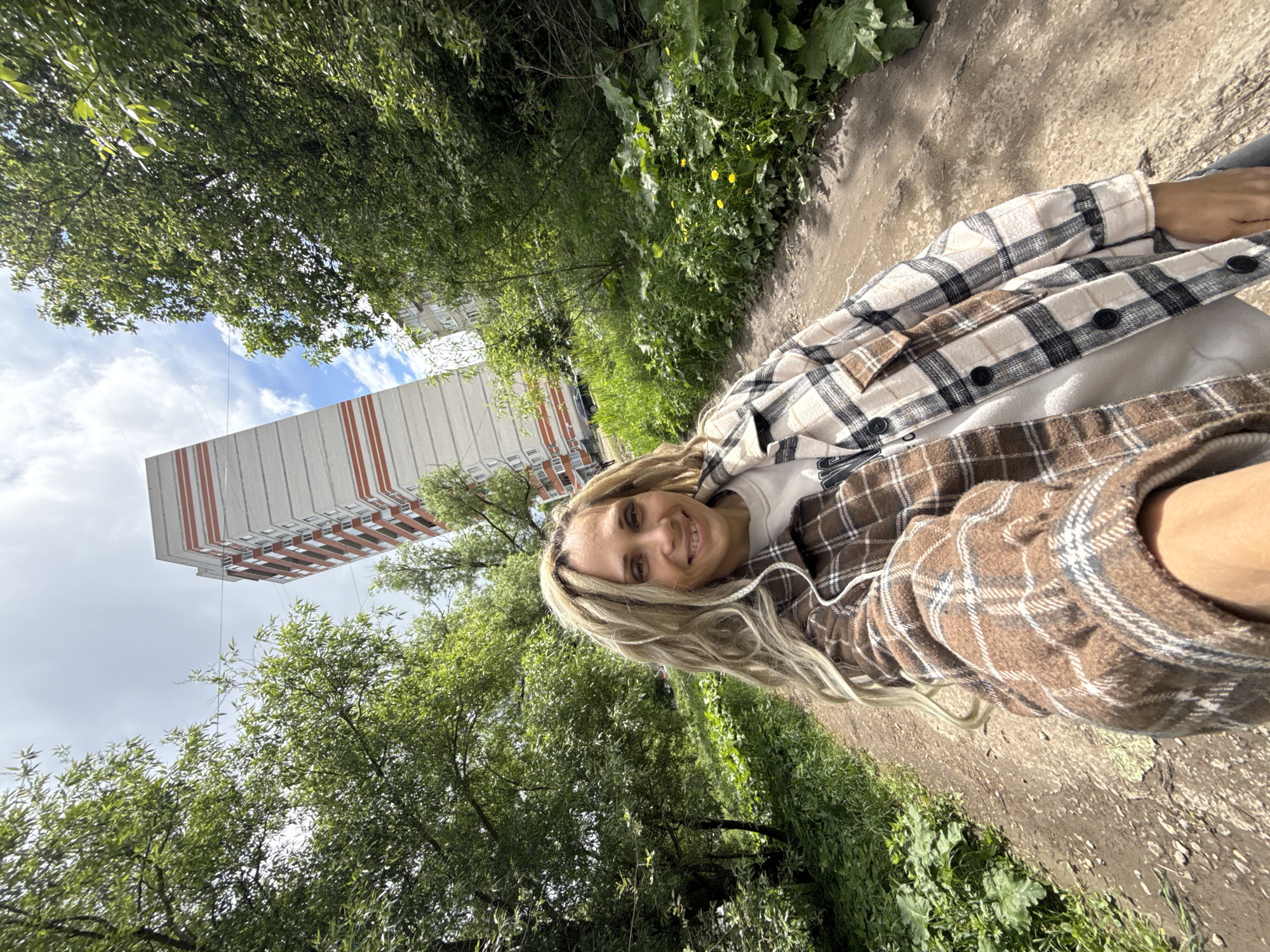 A young, smiling blonde woman is centered in a portrait-style photo taken outdoors in Rusanovo, Russia. She is wearing a white t-shirt under a light brown and white plaid shirt. The background consists of a sidewalk or path with green grass and trees on either side. In the distance is a tall, modern building with white and orange stripes. The sky is partially cloudy.