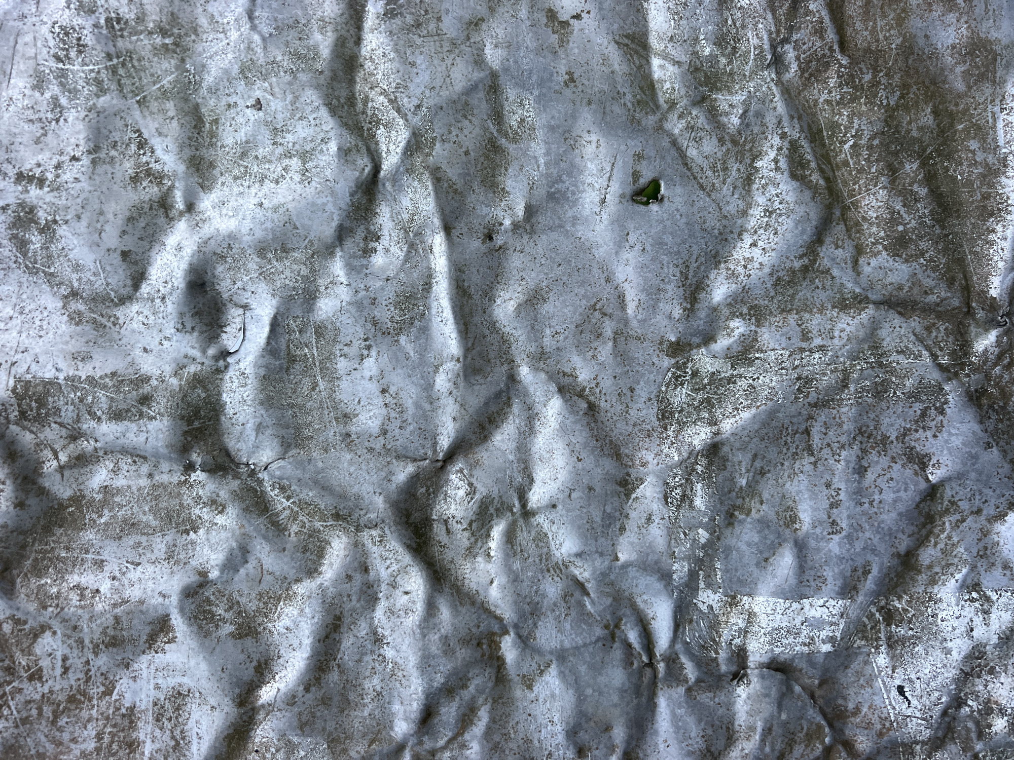 The image shows a close-up, full frame shot of a crumpled, weathered metal surface. The texture is rough and uneven, with visible dents, creases, and scratches. The metal appears to be a dull, silvery-gray color, with patches of darker brown and white discoloration, suggesting rust or paint wear. The lighting creates shadows and highlights that emphasize the three-dimensional quality of the crumpled surface. There are small specks of what might be dirt or debris scattered across the surface, and in one area, a small, bright green object is visible within a tiny hole or indentation. The overall impression is of a damaged and aged material, likely exposed to the elements. The location is Unknown, Unknown.